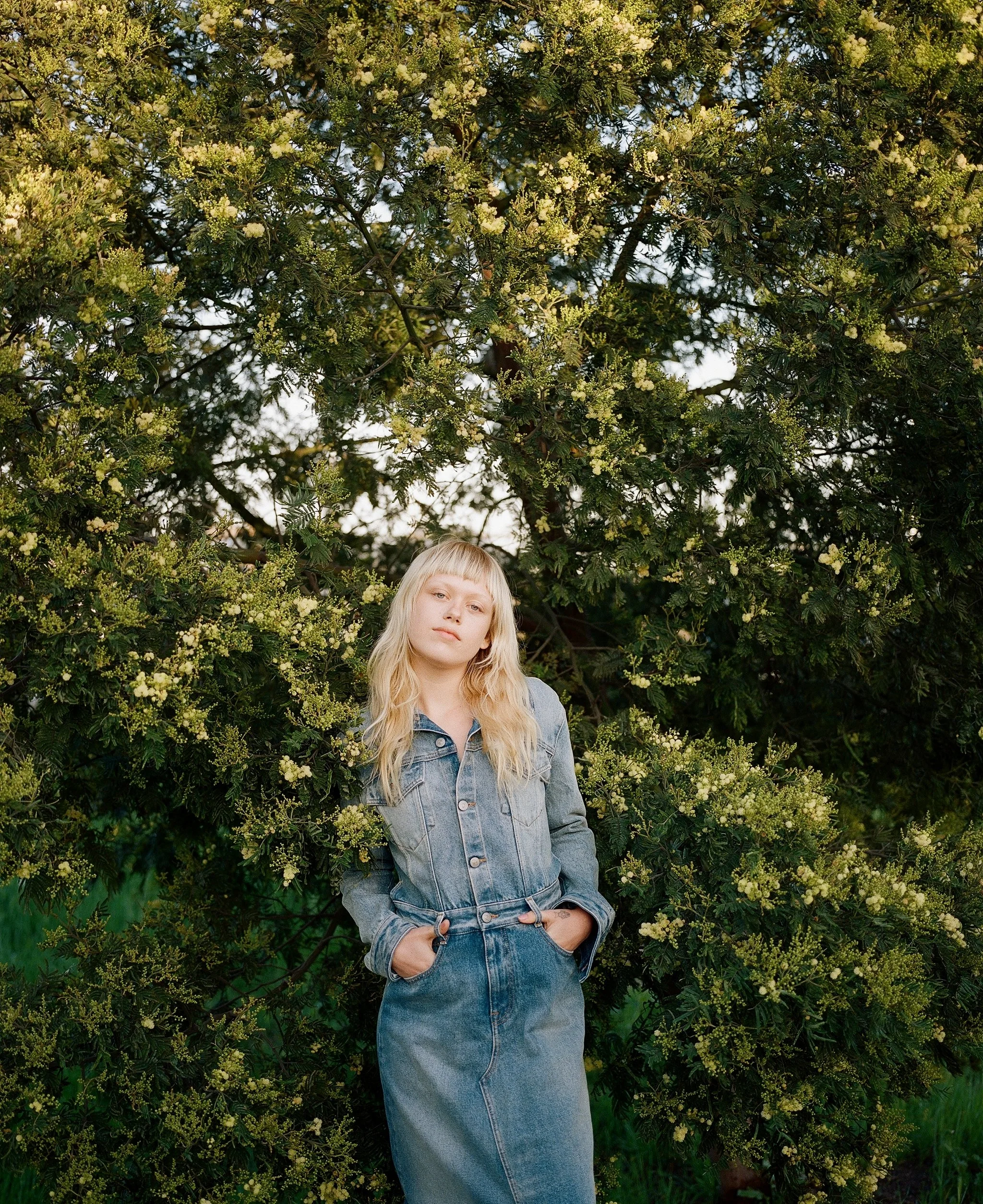 Medium format editorial film photograph — blonde woman in denim maxi dress standing among wildflowers beside highway embankment, direct gaze, square format, by Radostina Boseva