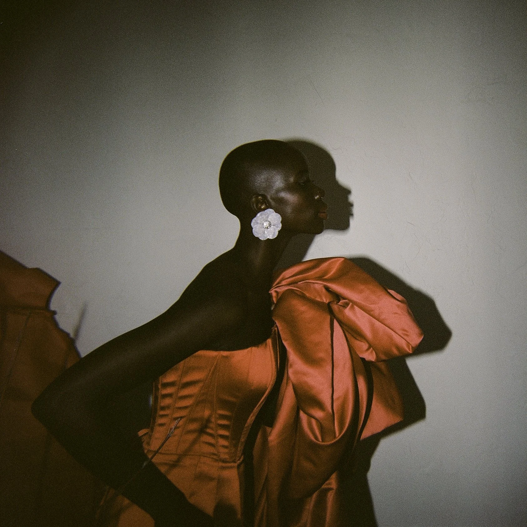 Editorial film portrait — woman in sculptural red satin gown in profile, her shadow cast on the wall behind her, white flower earring, by Radostina Boseva