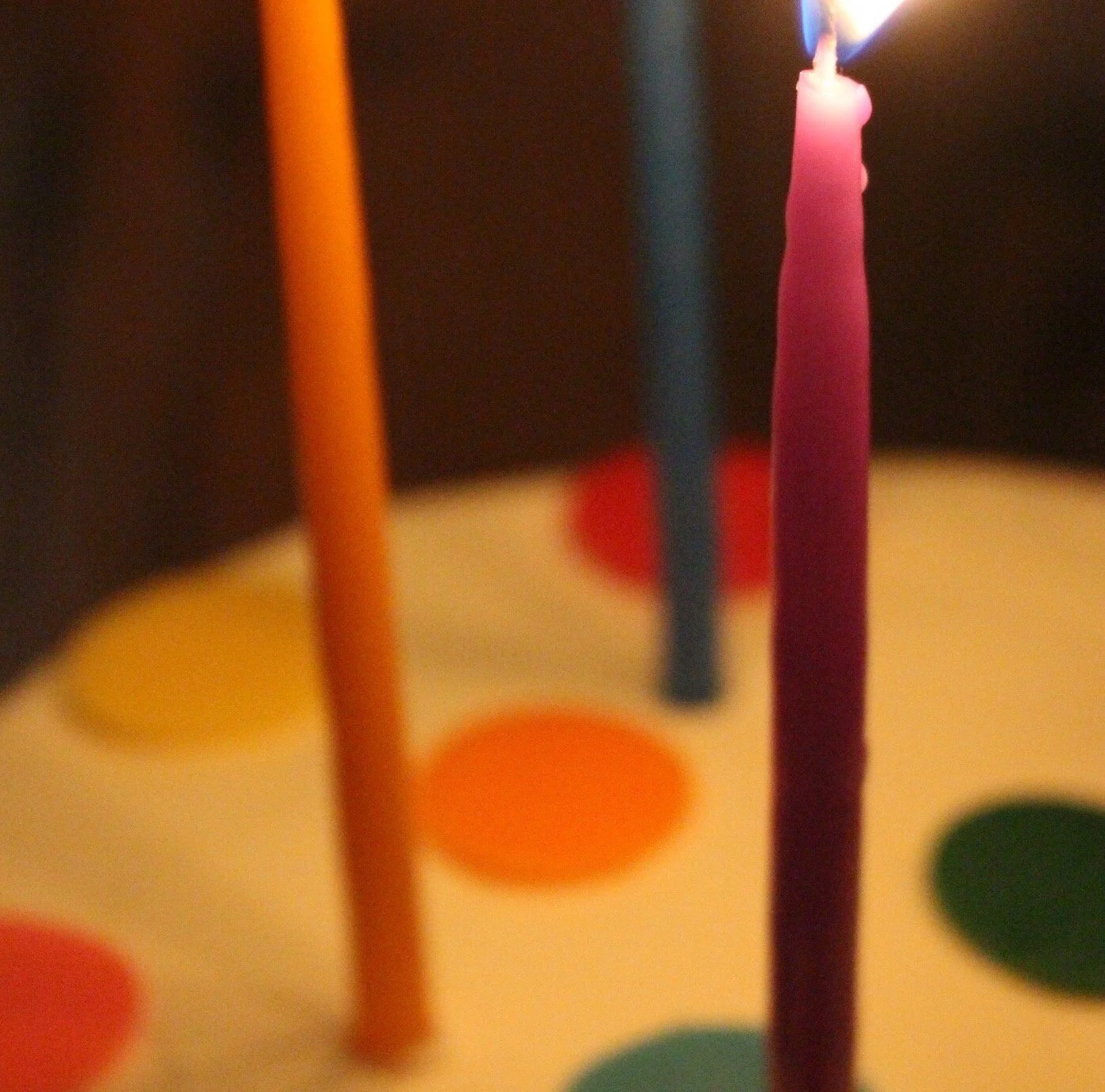 Close-up of a birthday cake with three lit candles in orange, pink, and blue, with multicolored polka dots on the cake's white surface.