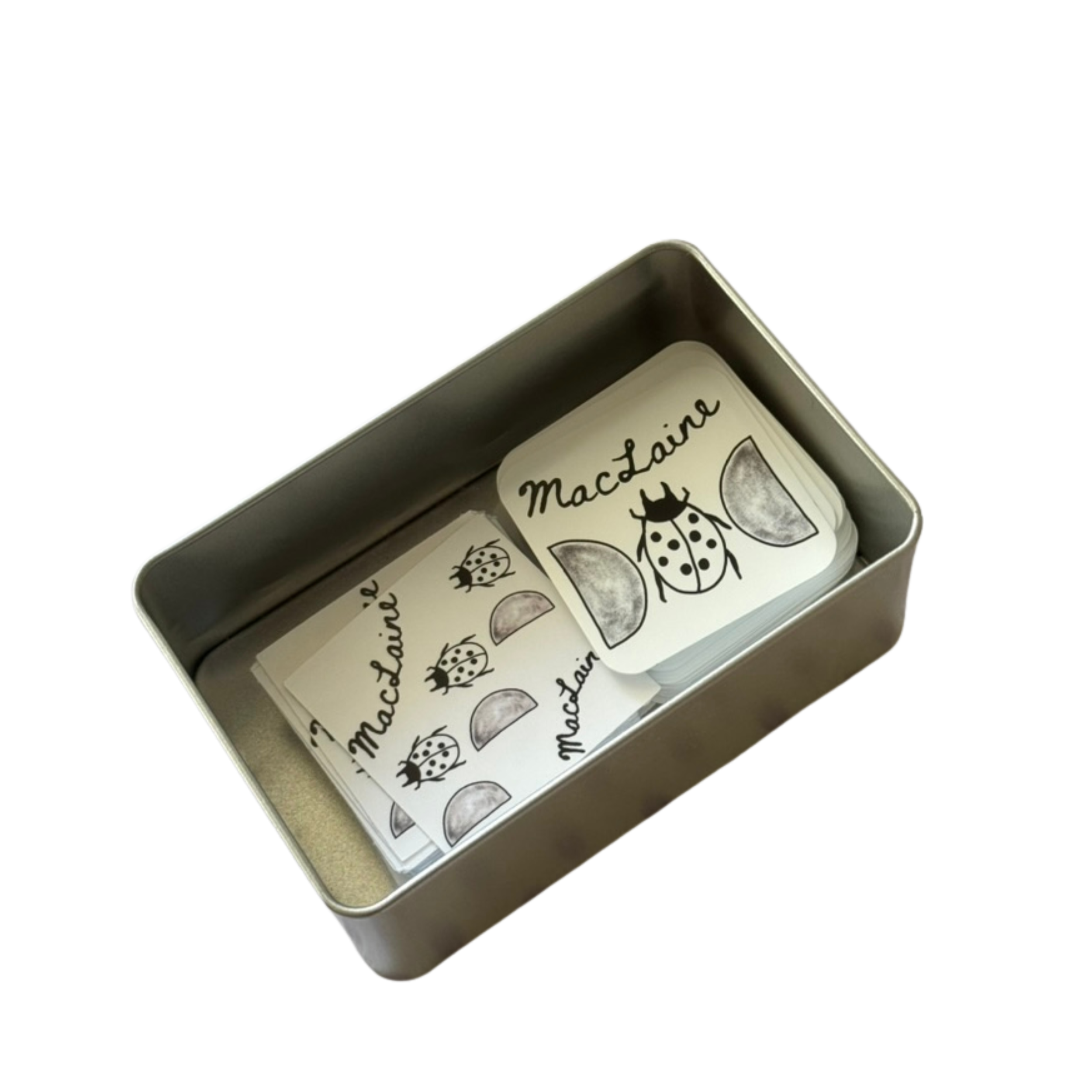 Stack of cards with ladybug and half-moon designs, labeled 'Macaqine' in a tin box.