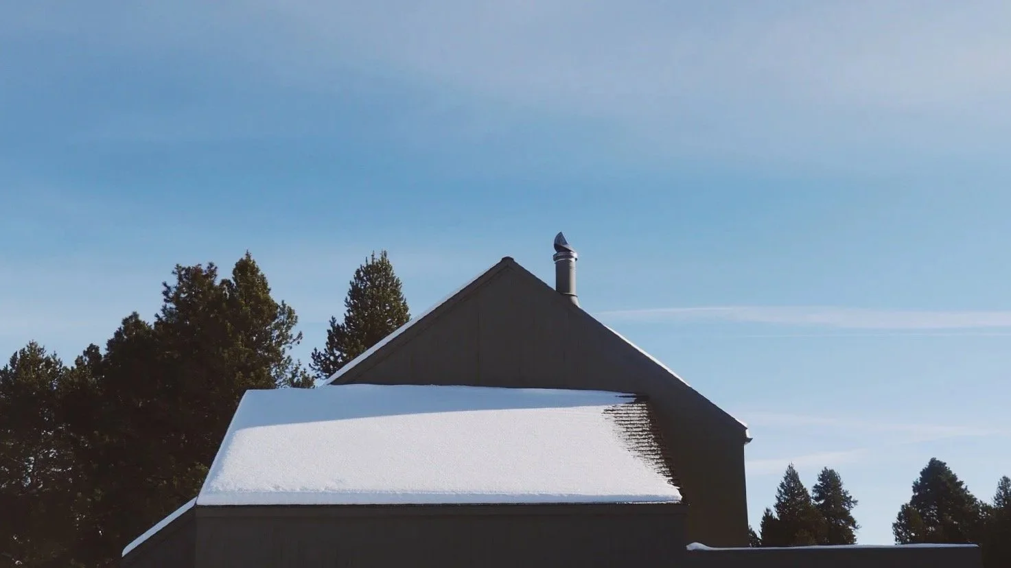 A house with a sloped roof covered in snow, surrounded by tall evergreen trees under a clear blue sky.