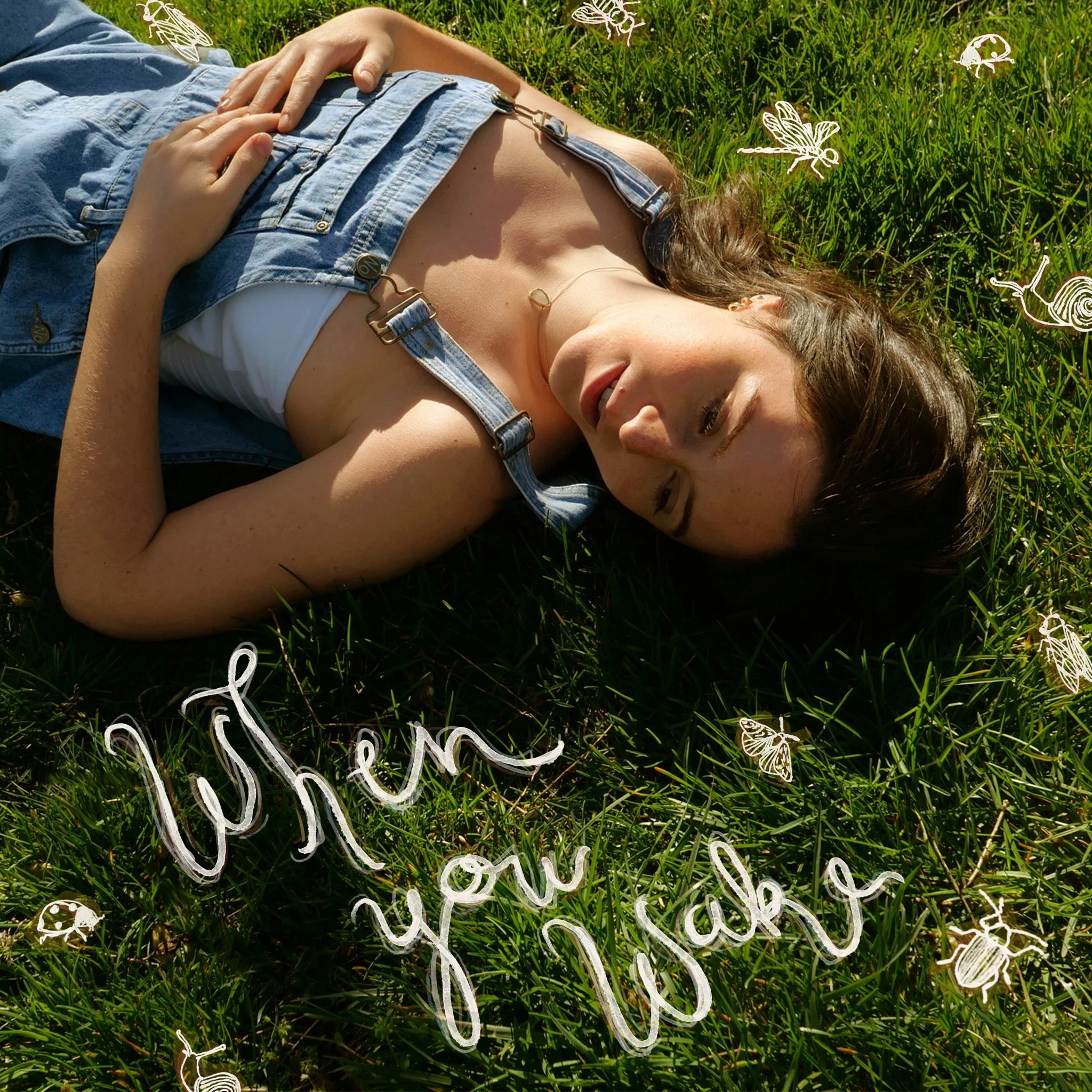 A young woman lying on a grassy field with her eyes closed, wearing denim overalls and a white top. The photo includes white doodles of insects and the handwritten phrase "when you ask" overlayed on the grass.