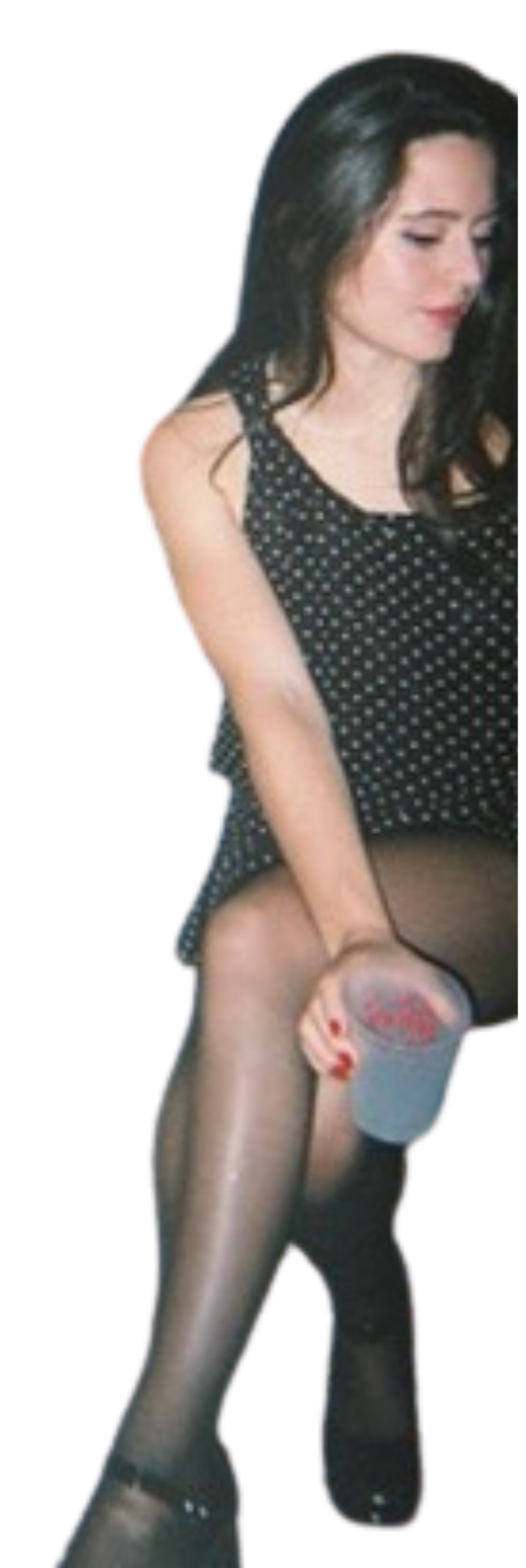 A woman with long dark hair wearing a polka dot dress and black stockings, holding a plastic cup with a beverage, against a black background.