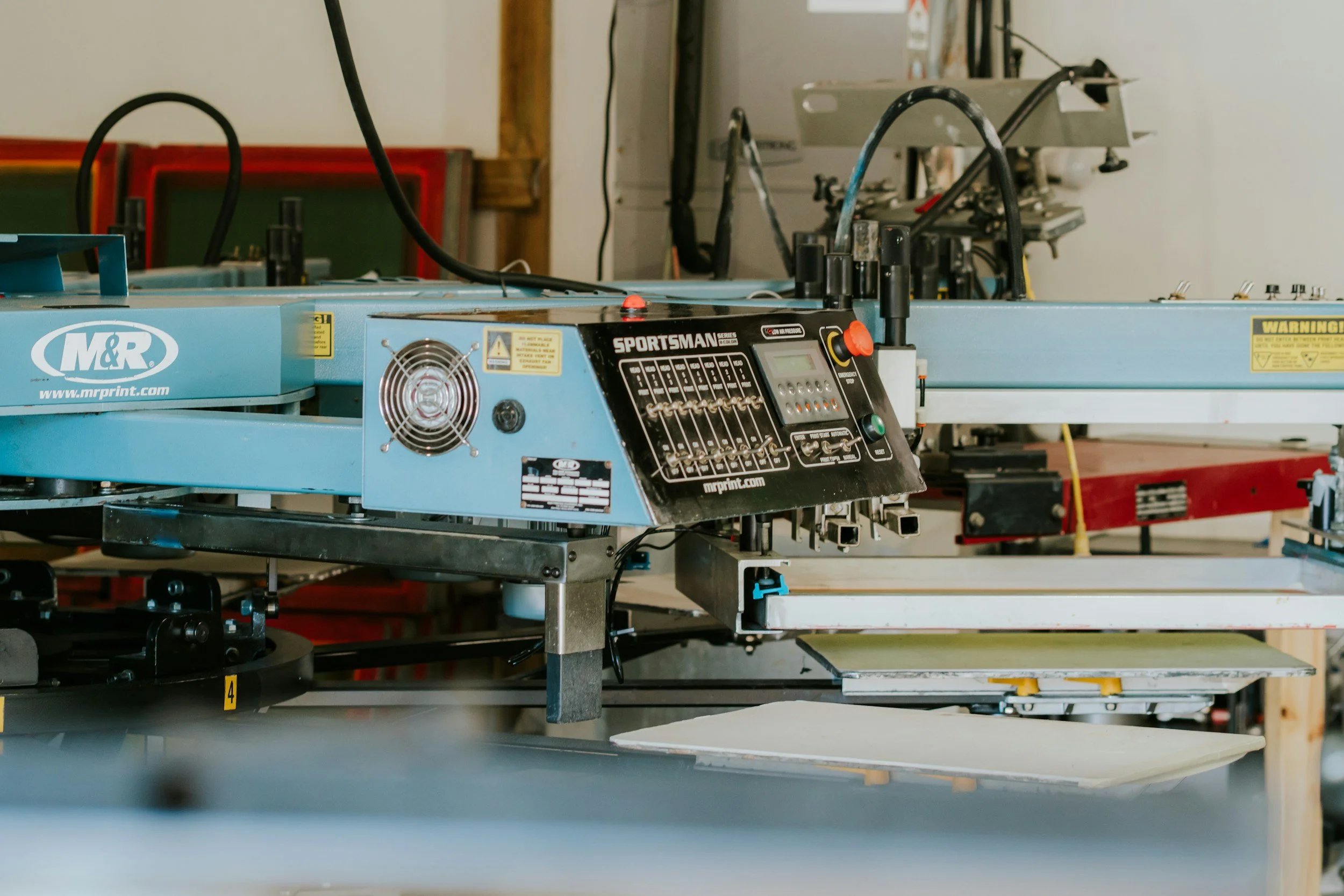 A blue, industrial machine with controls labeled "SPORTSMAN" and "M&R Print" in a workshop. In the background, there are workbenches and tools, and an unfinished metal frame is mounted on the machine.