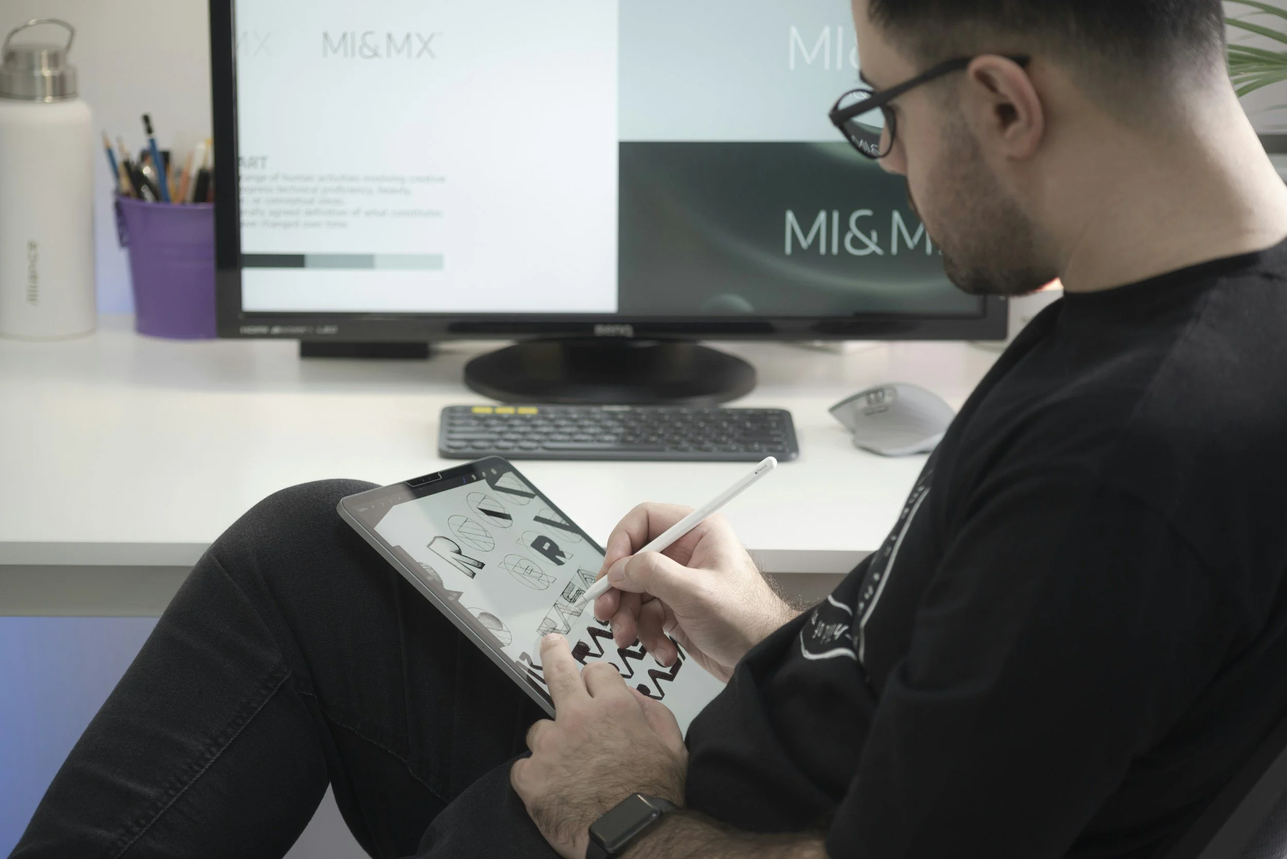 Man with glasses sitting at a desk, using a stylus on a tablet. The desk has a computer monitor, a keyboard, a mouse, and a purple pencil holder with pens and pencils. The monitor displays the label 'MI&MX'.