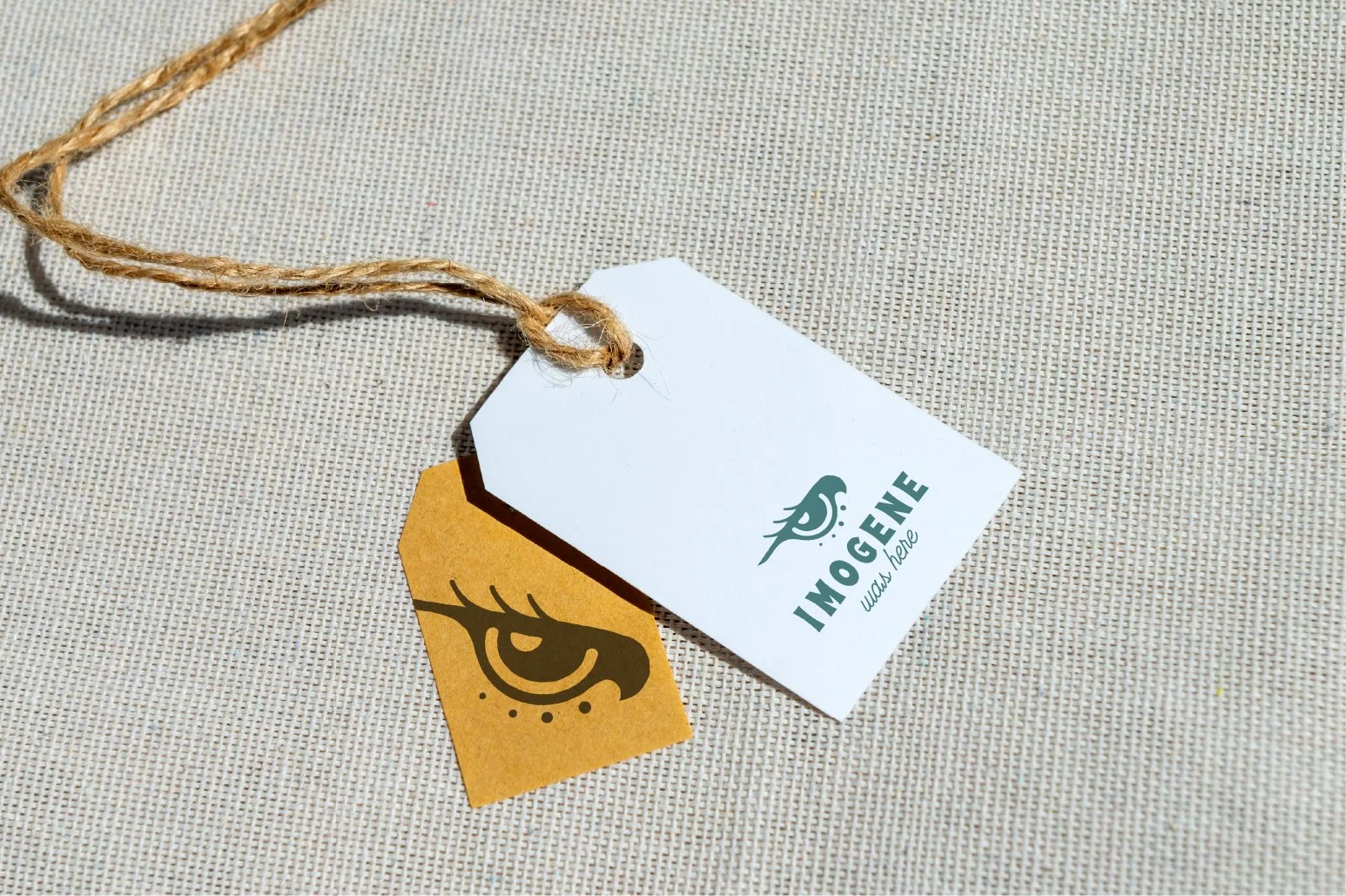 Two tags attached to a piece of twine lying on a textured beige fabric background. One tag is white and has a stylized bird logo with the text "IMOGENE" and "wild & free" beneath it. The other tag is yellow and features a black illustration of an eye