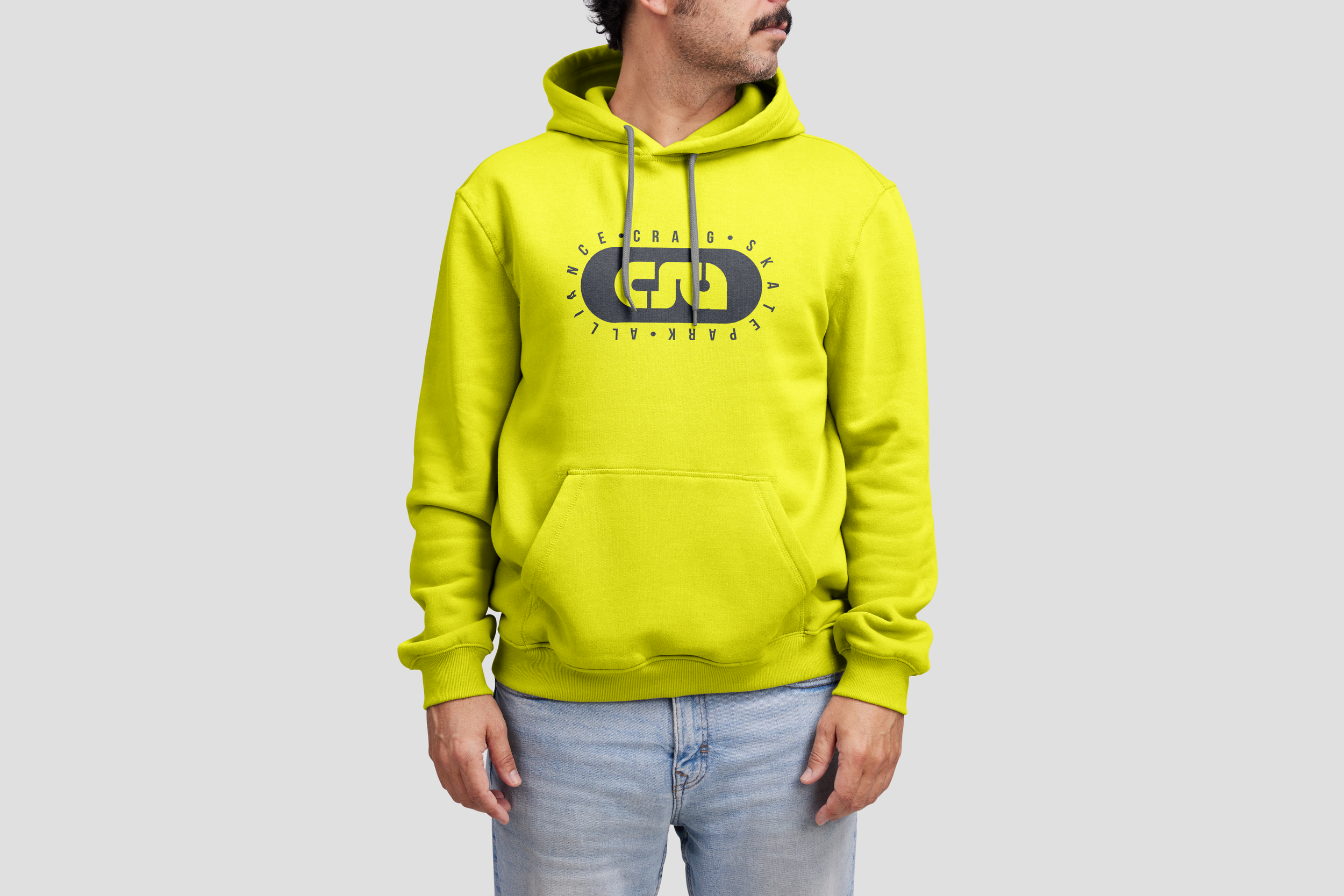 A man is wearing a bright yellow hoodie with a logo and text on the front, and light blue jeans, standing against a plain light gray background.