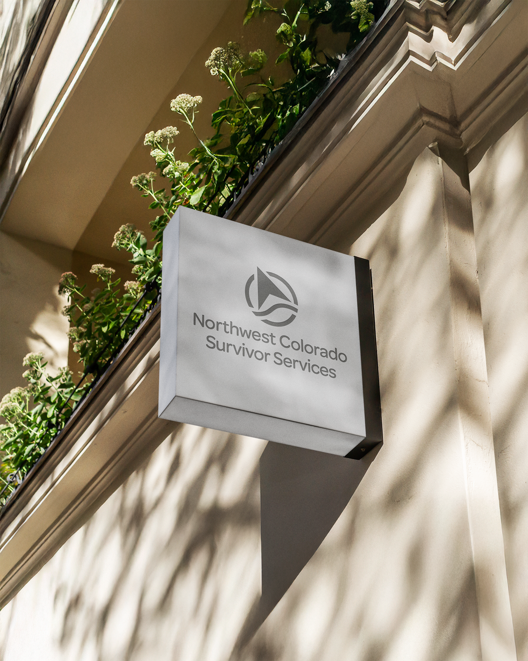 Square sign on a building with the logo and text 'Northwest Colorado Survivor Services', a window with plants on the ledge above, and sunlight casting shadows on the wall.