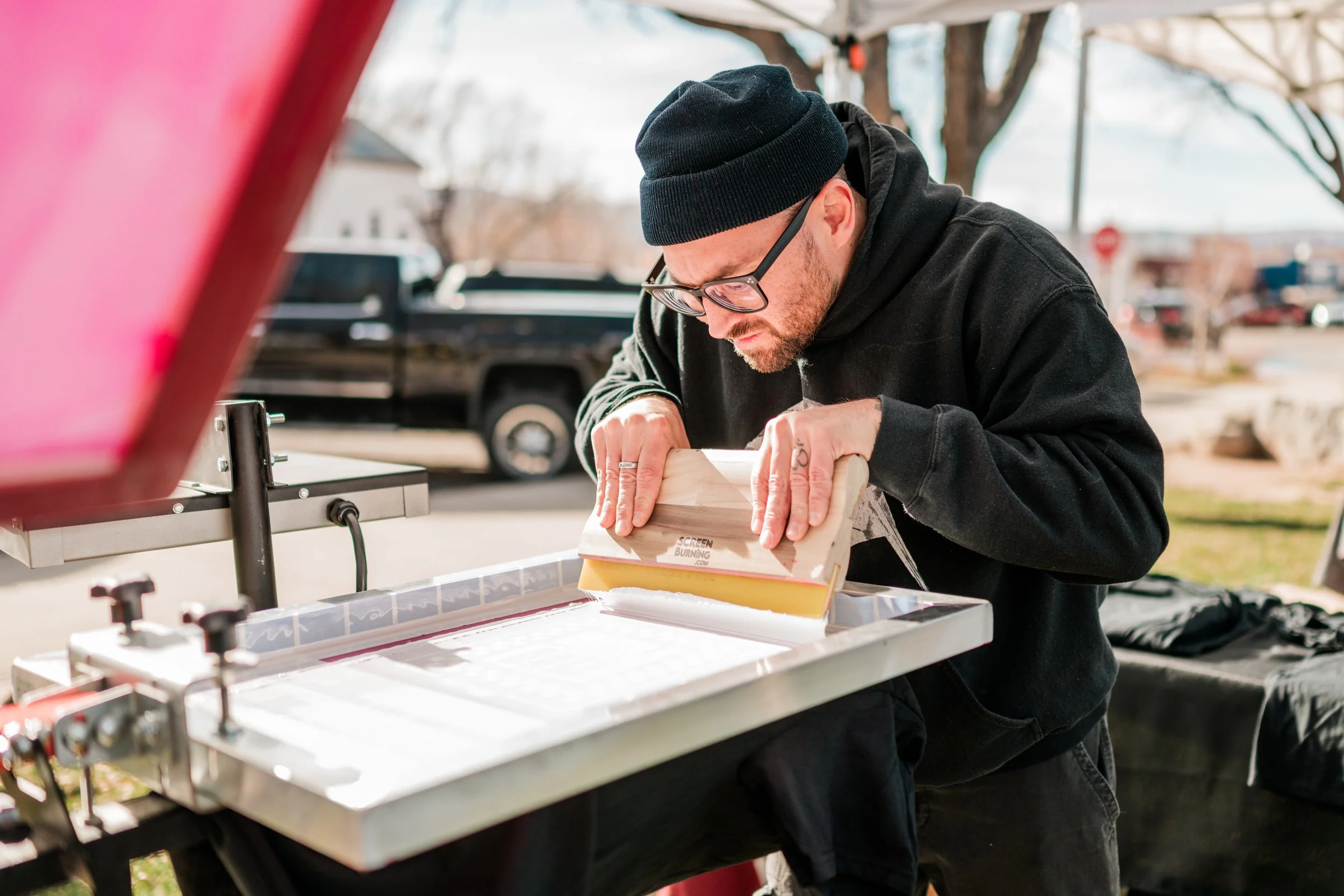 A man printing t-shirts using a heat press machine outdoors. He is wearing glasses, a black beanie, a black hoodie, and focused on the task.