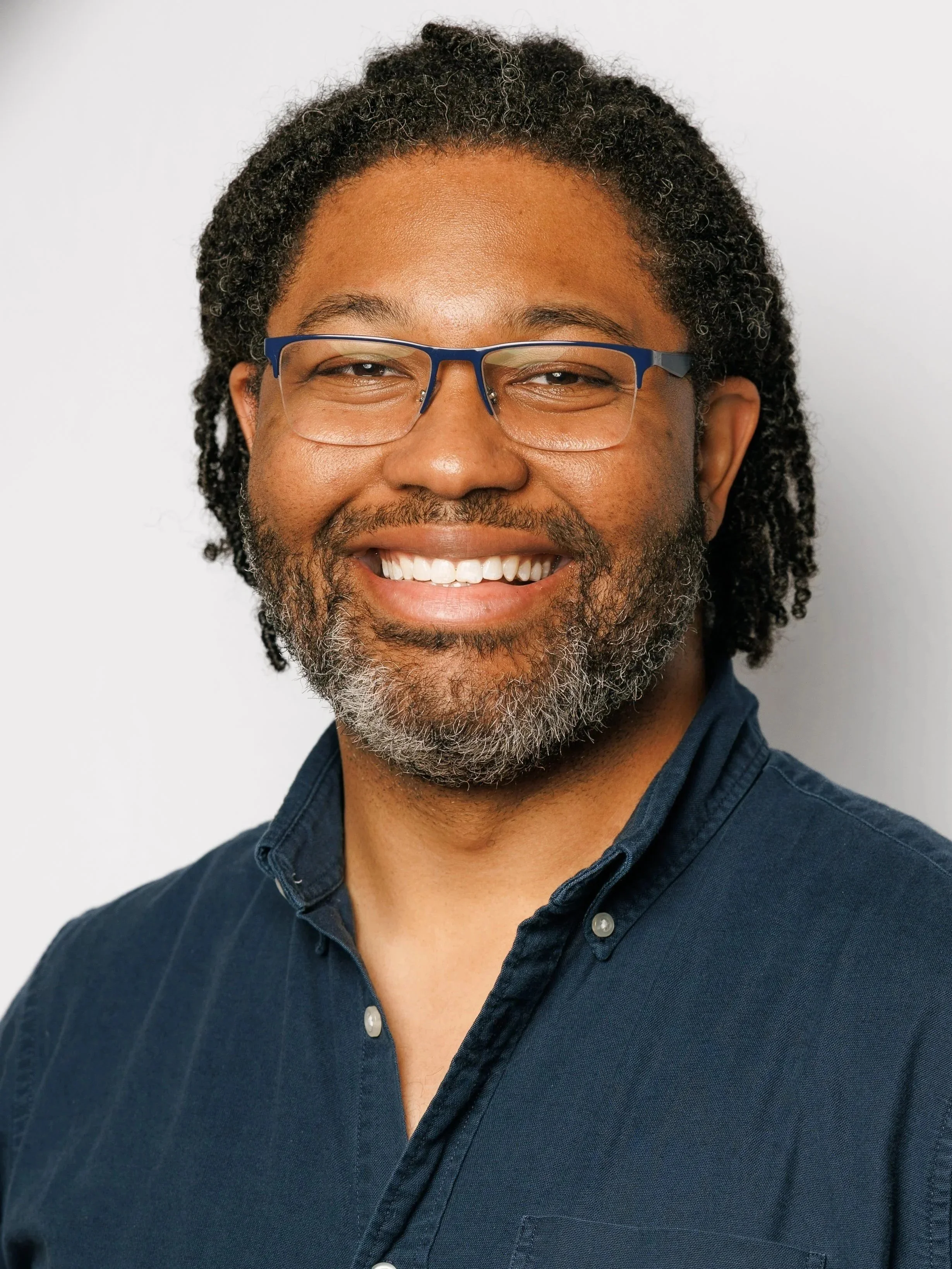 A smiling man with glasses, a beard, and dreadlocks poses in front of a backdrop with the logo of Ascend Music Studios. He is wearing a navy blue polo shirt and jeans.