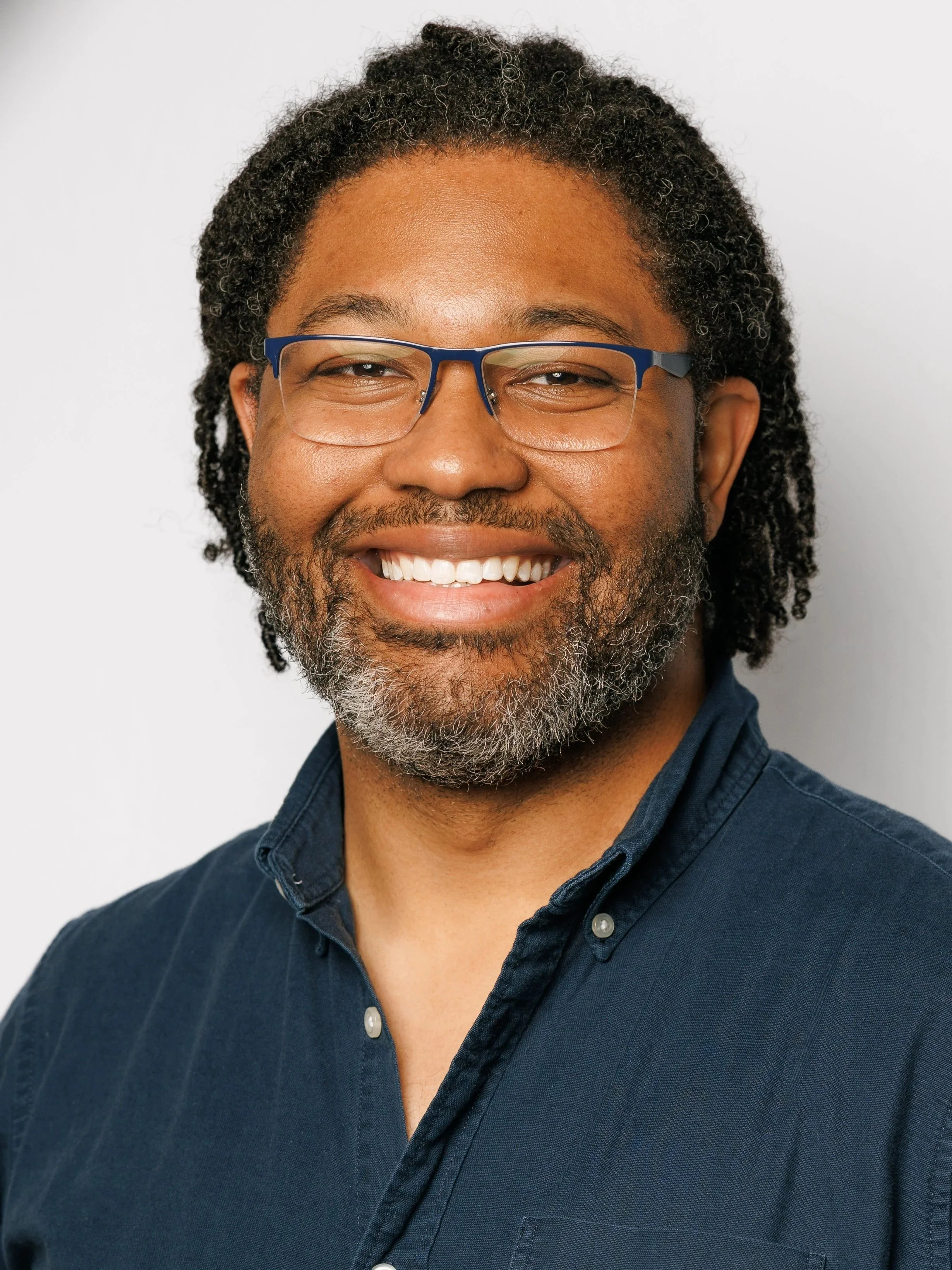 A man with black curly hair, glasses, and a beard, smiling, wearing a dark blue polo shirt with a small logo on the left chest, standing in front of a white backdrop with red and blue logos for Ascend Music Studios, with his right hand in his pocket and left hand resting on his hip.