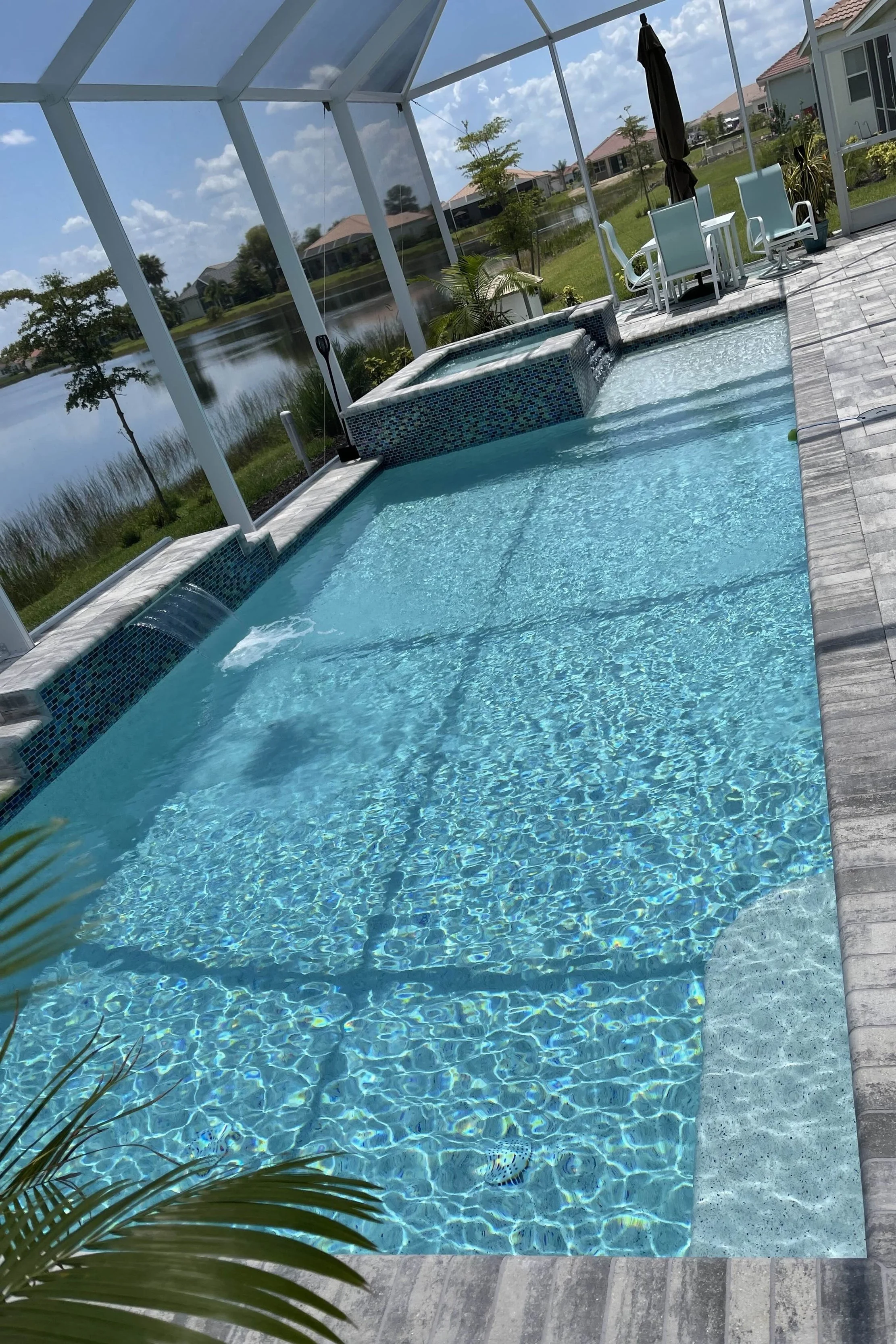 Backyard swimming pool with hot tub, enclosed by a screened structure, overlooking a lake with houses, chairs, and a table with an umbrella