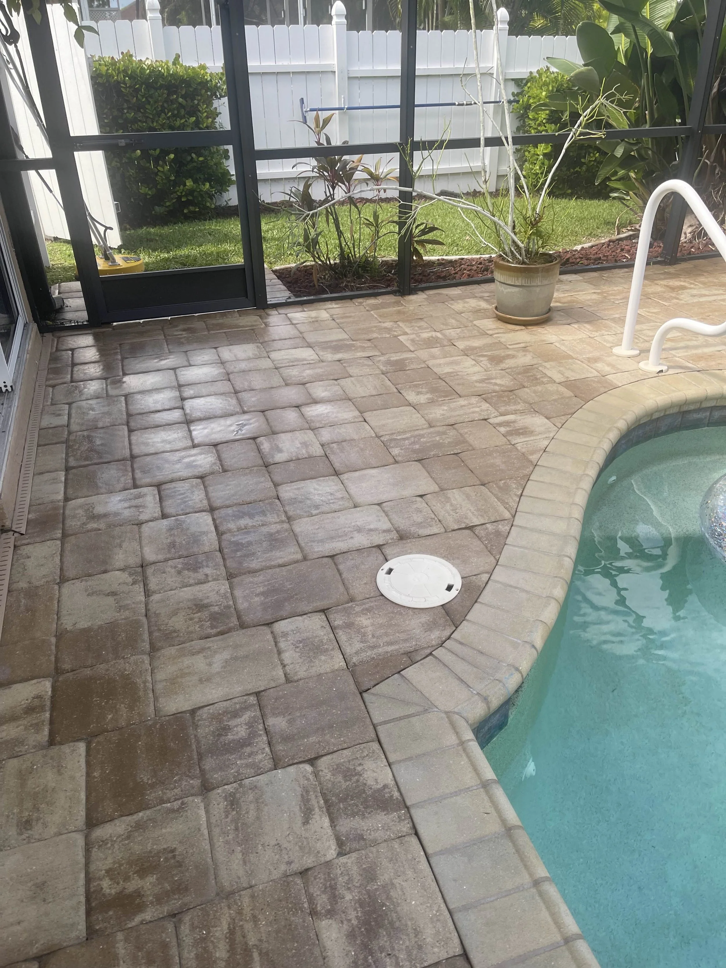 Enclosed backyard patio with stone pavers, a swimming pool with a curved edge, potted plants, and a white fence.