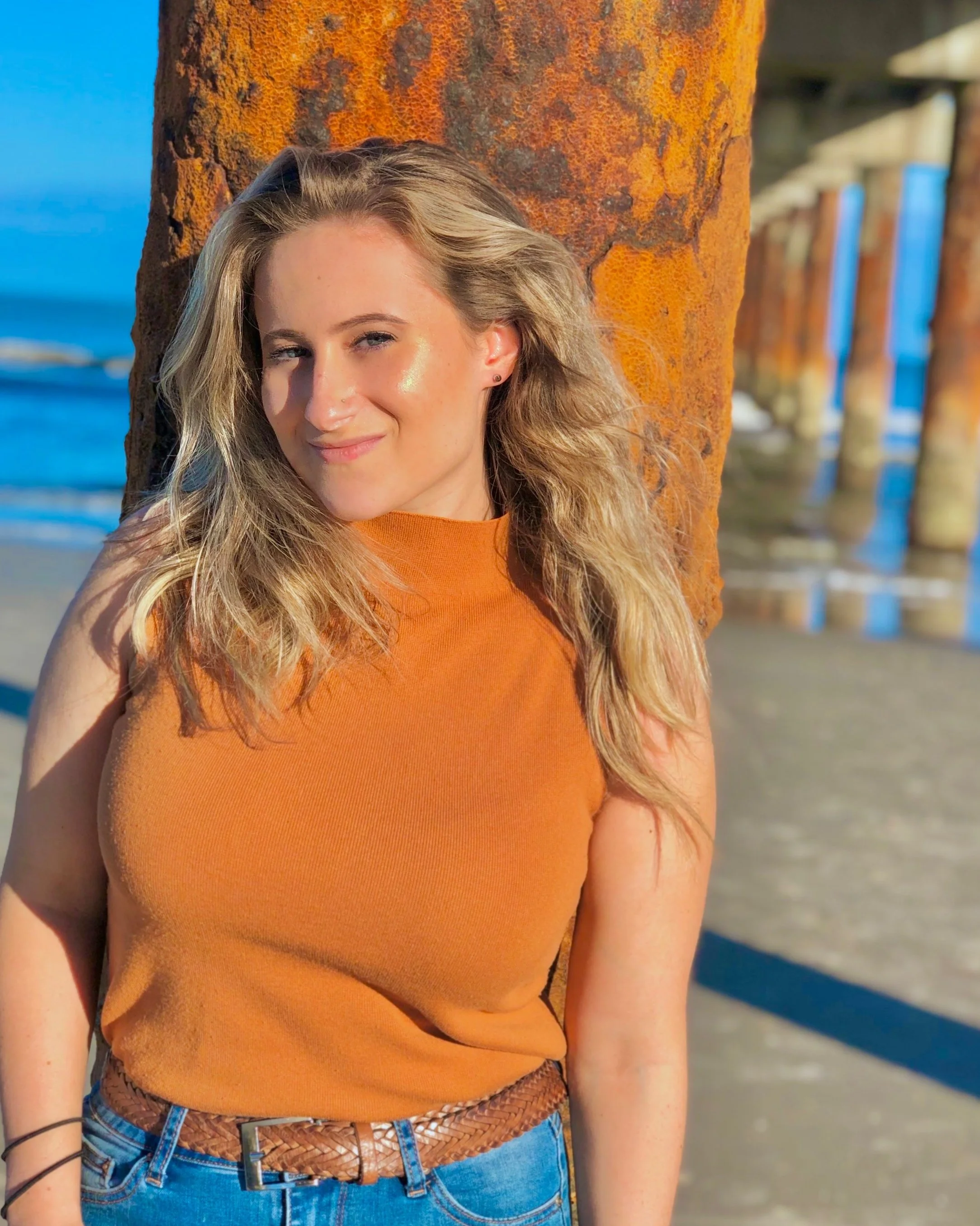 A blonde woman in an orange sleeveless top and jeans leaning against a rusty pier post at the beach during sunset.