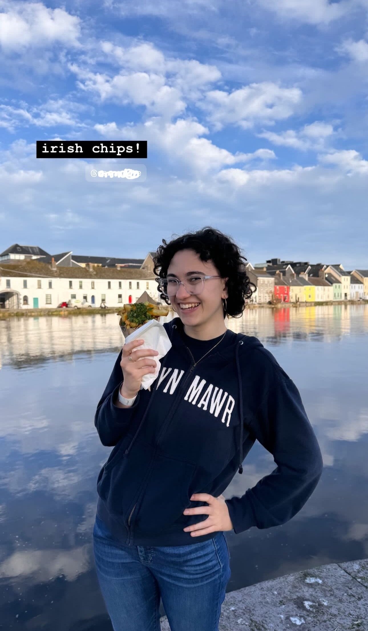 A young woman with curly black hair and glasses smiling as she holds a cone of Irish chips near a river with reflective water, colorful houses in the background, under a partly cloudy sky.