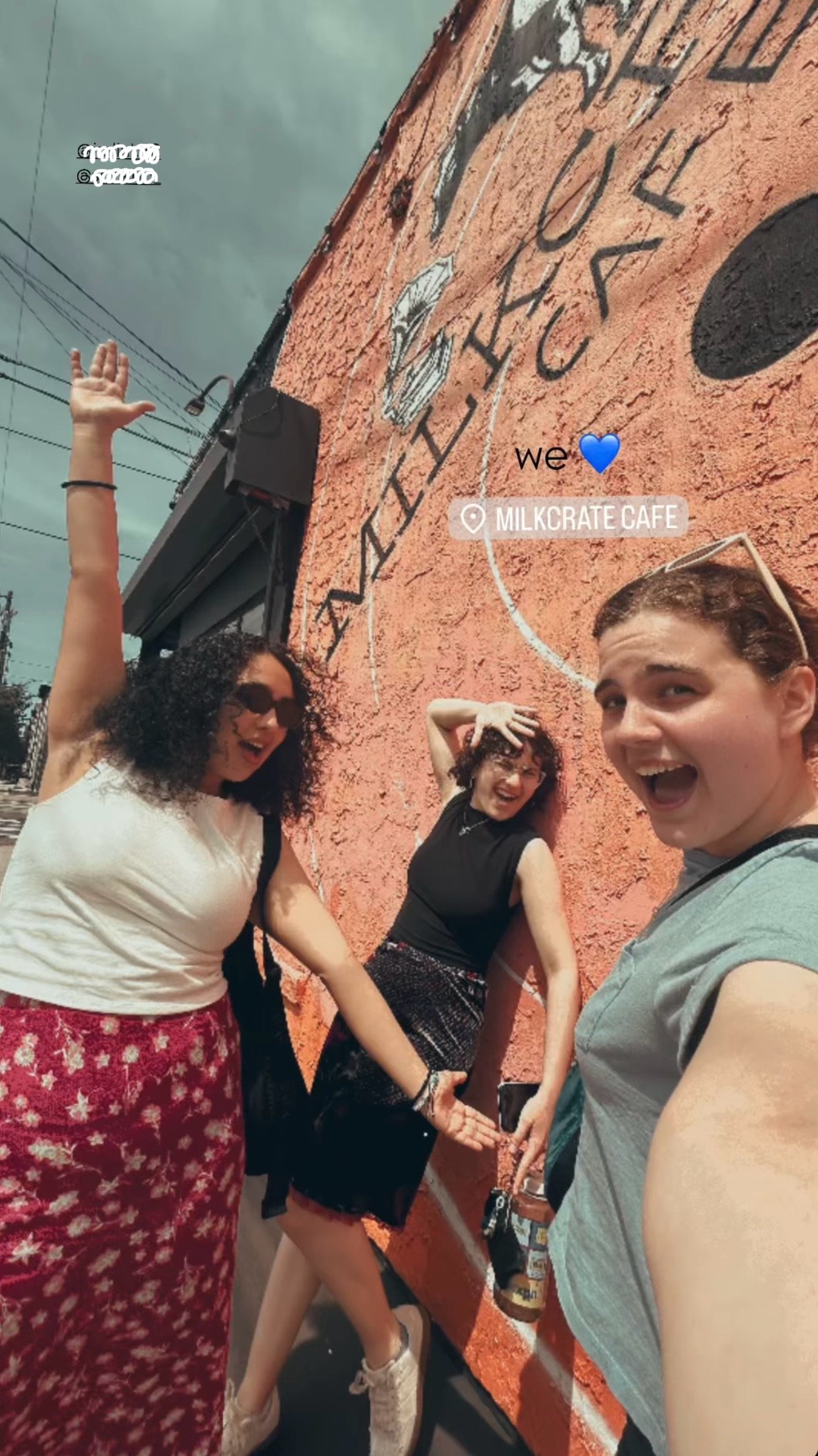 Three women are smiling and posing in front of an orange wall with black lettering at Milkcrate Cafe, one woman raising her arm, another touching the wall, all enjoying their time together.