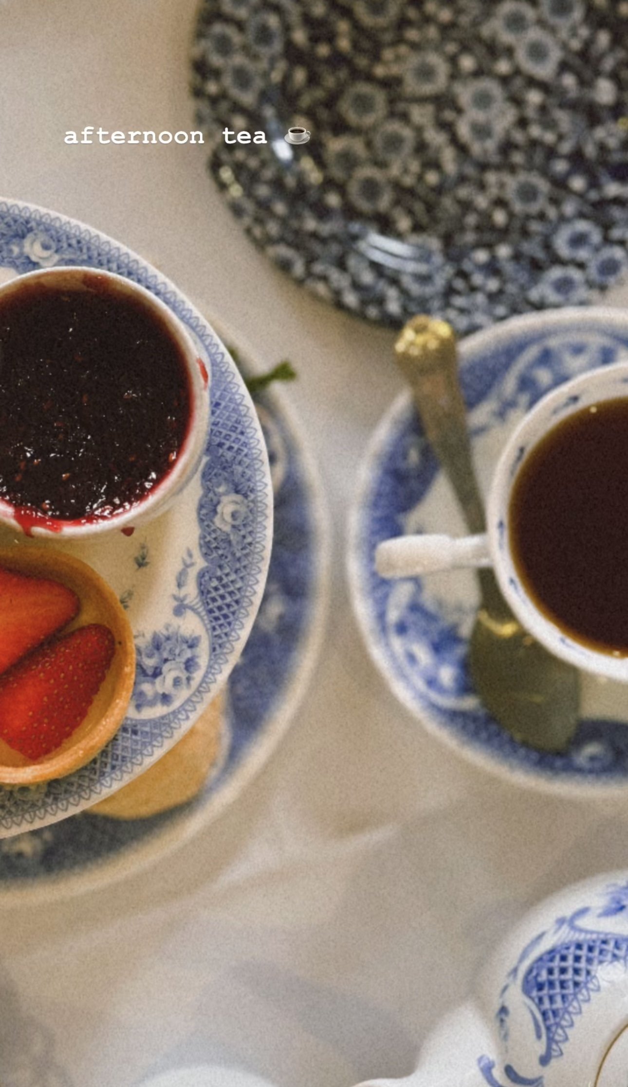 Two cups of tea on matching blue and white saucers with a small bowl of strawberry jam and a tart with strawberries, on a white table with a patterned cloth in the background.