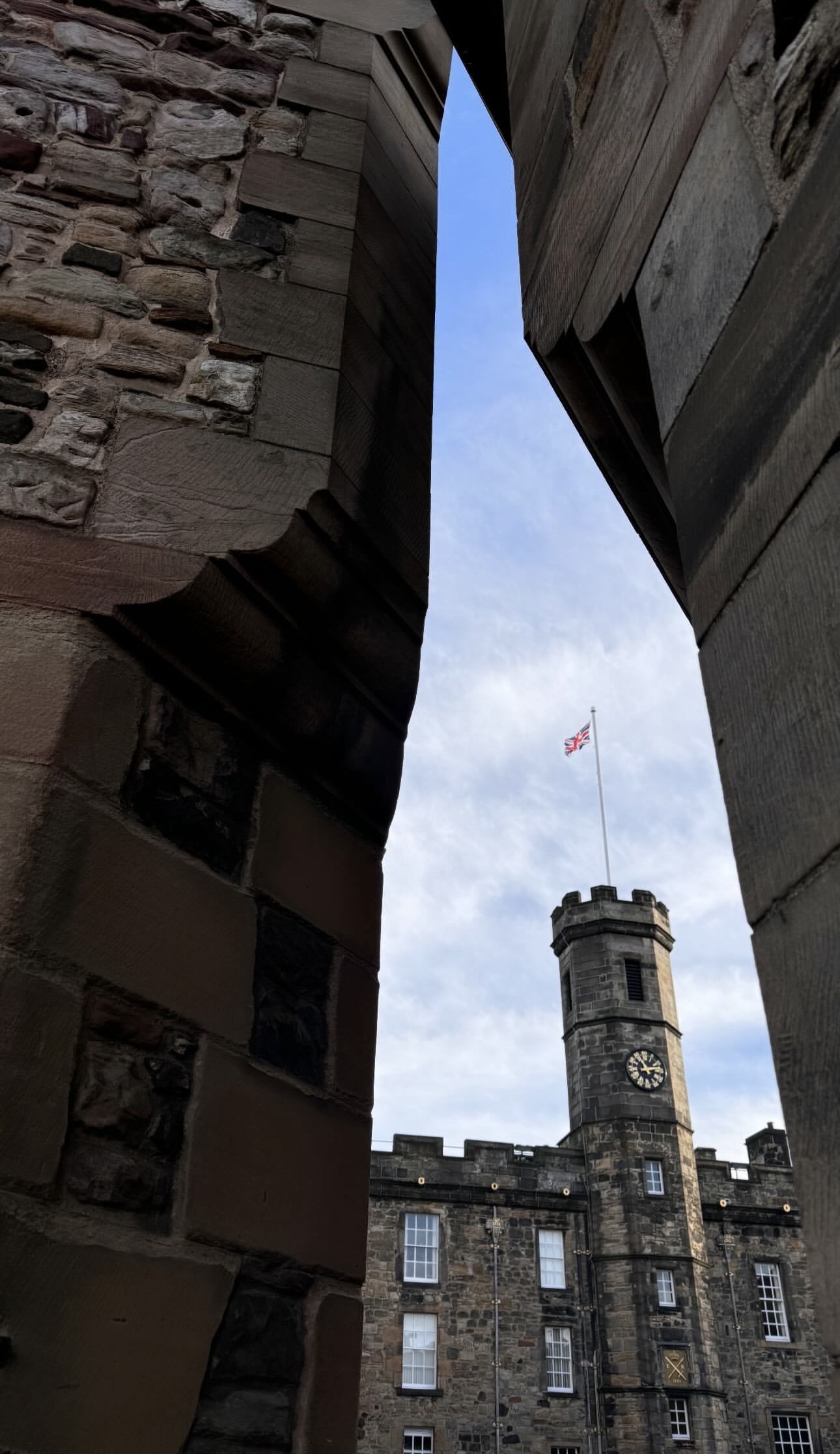 Low-angle view of a historic stone building with a clock tower and a flag on top, framed between brick and wooden walls in the foreground.