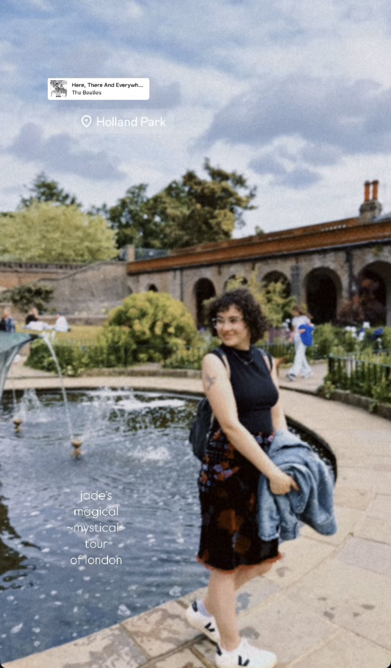 A woman with curly hair and glasses, standing by a fountain in a park, holding a jacket, with garden and brick building background.