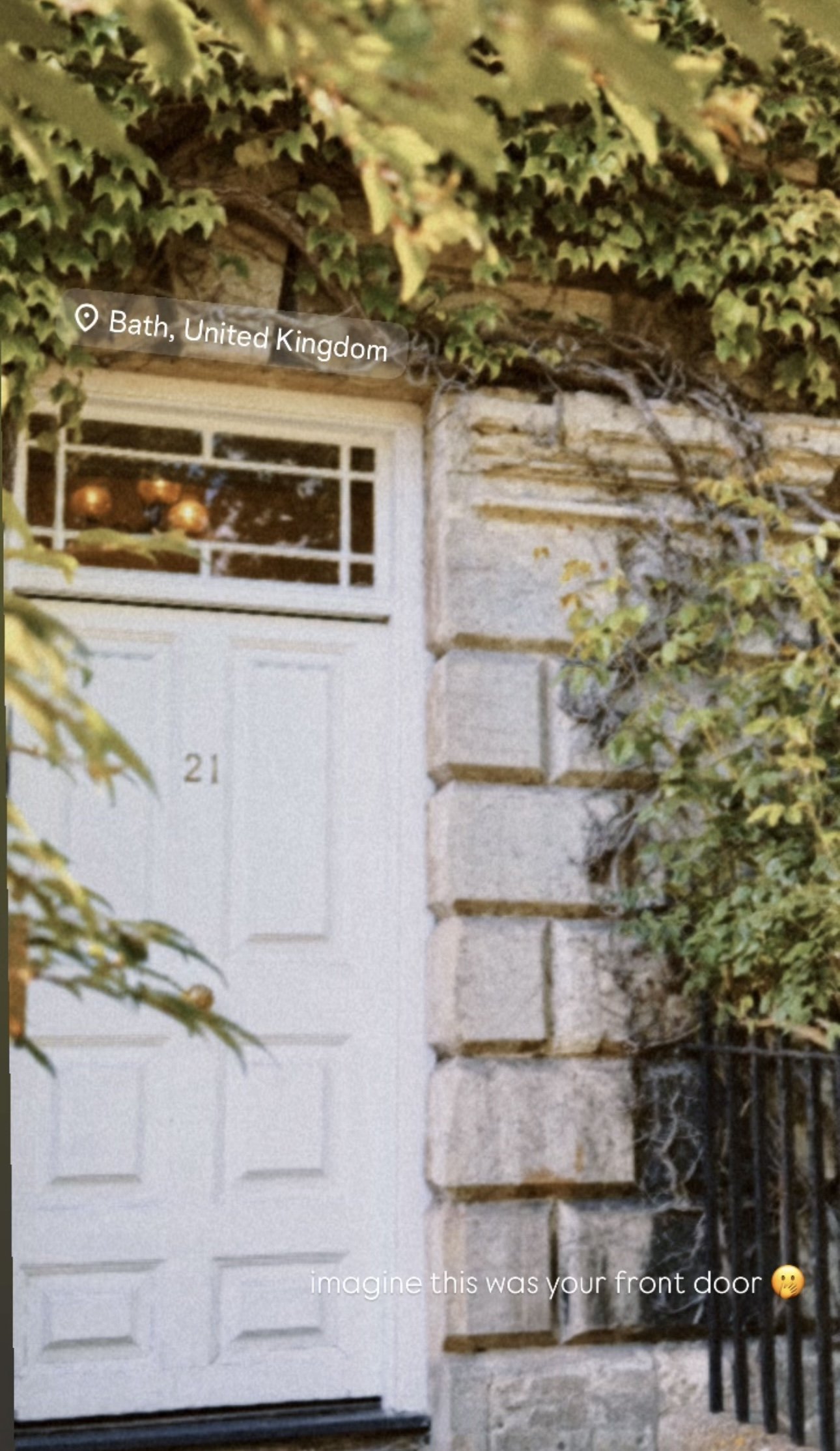 A white front door with the number 21, surrounded by ivy and greenery, in Bath, United Kingdom. There are decorative items visible through the window, and a caption suggesting imagining it as your front door.