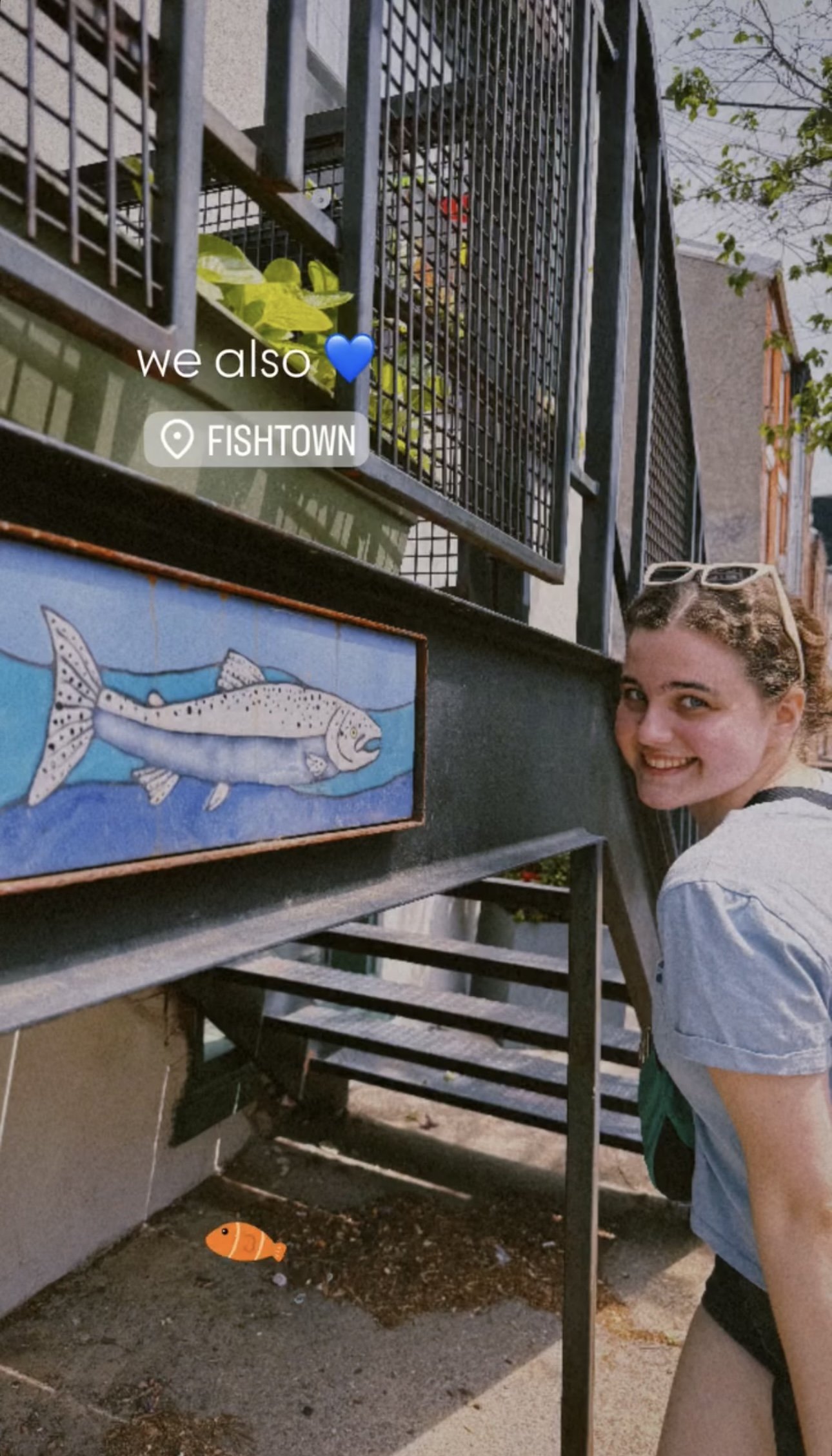 A smiling woman with sunglasses on her head leaning beside a mural of a fish on a street in Fishtown. The background shows residential buildings and a metal staircase.