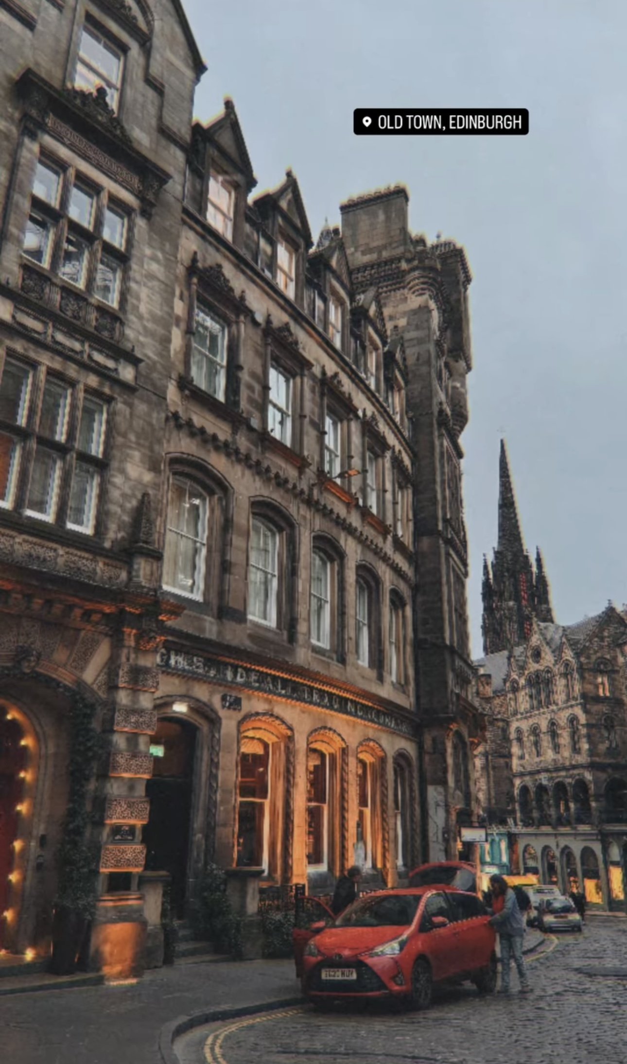 Historic stone buildings in Edinburgh’s Old Town with cars parked on cobblestone street and people standing near a red car.