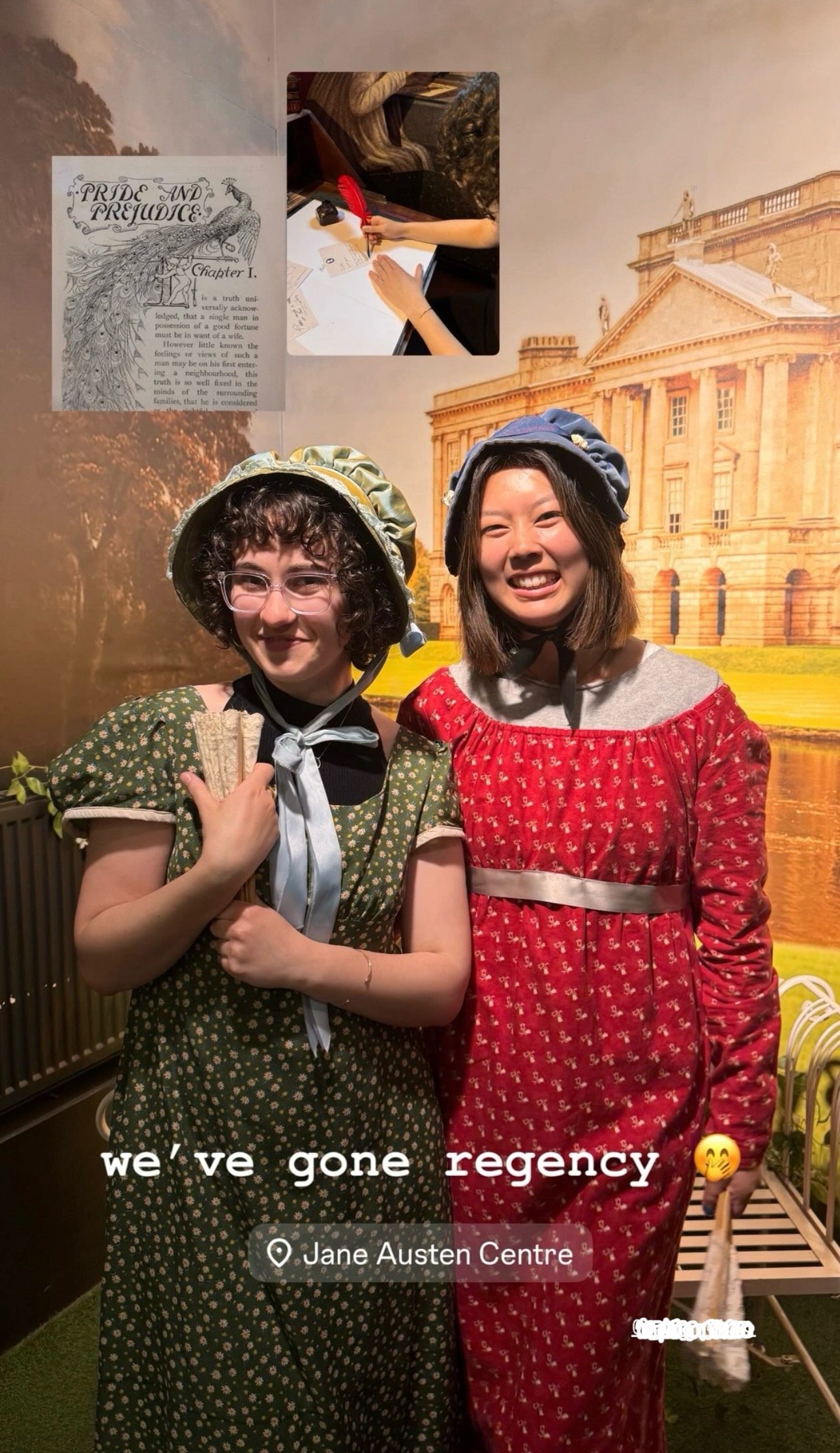 Two women in period costumes standing in front of a large backdrop of a historic building, smiling at the camera. One woman is wearing a green dress with white floral pattern and a bonnet, holding a small prop, while the other wears a red dress with