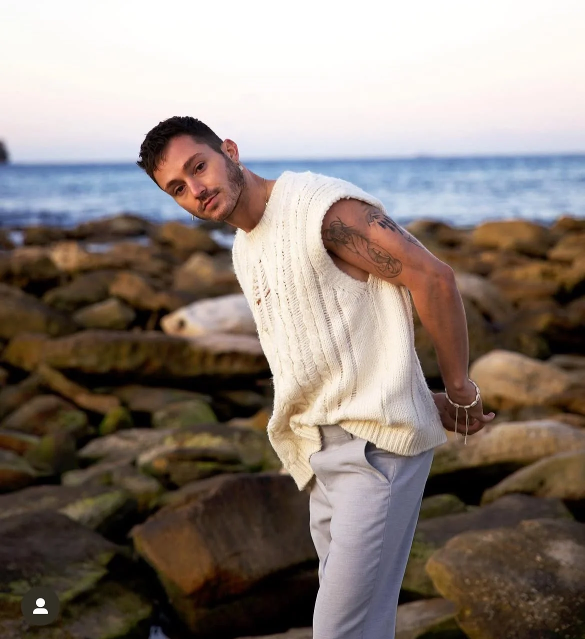 A man with dark hair, a beard, and tattoos on his arm stands on rocks by the sea, wearing a sleeveless white knit vest and light-colored pants, looking at the camera with a slight smile.