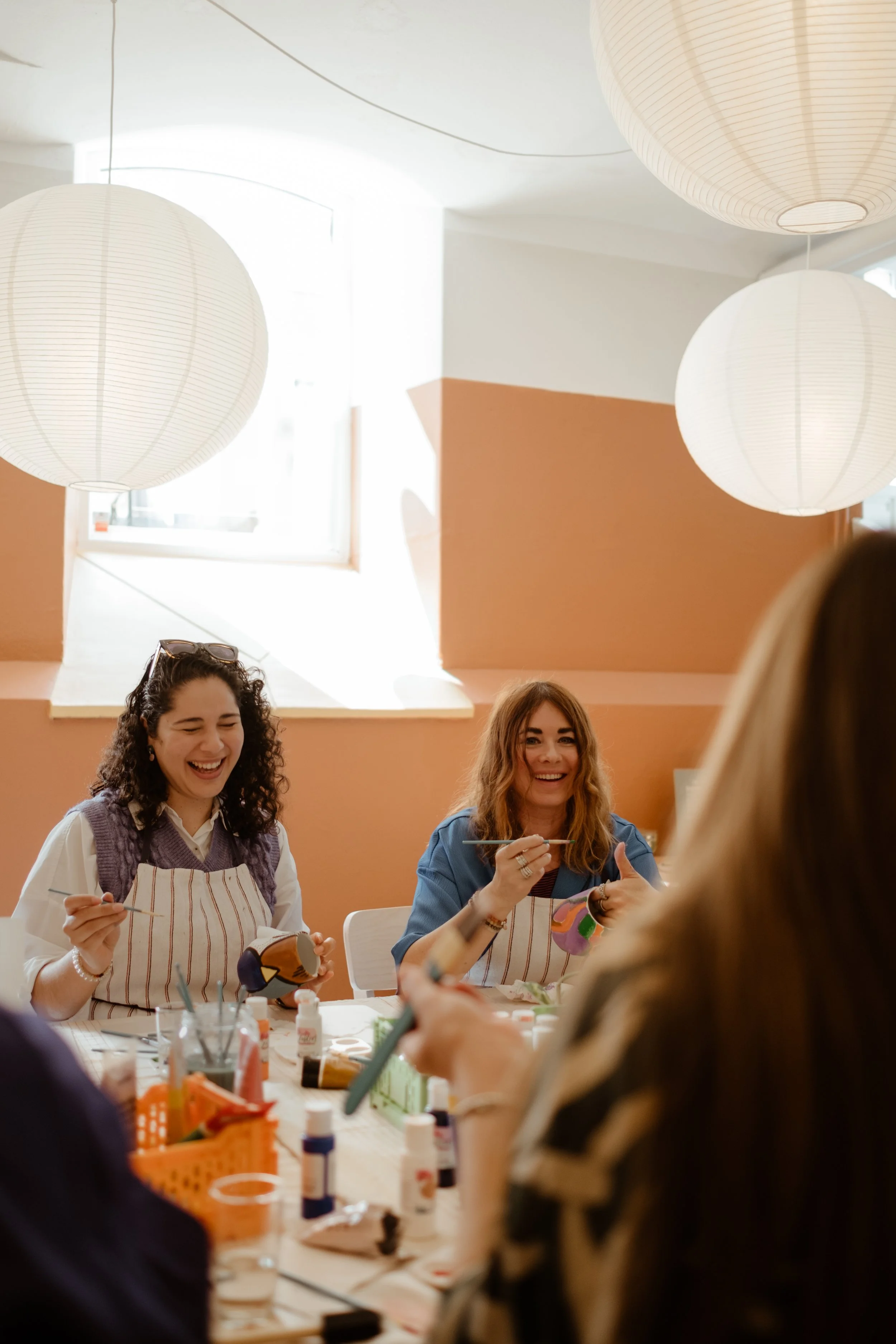 Women participating in a painting or arts and crafts activity, sitting at a table with art supplies, smiling and enjoying themselves in a well-lit room with large paper lanterns hanging from the ceiling.