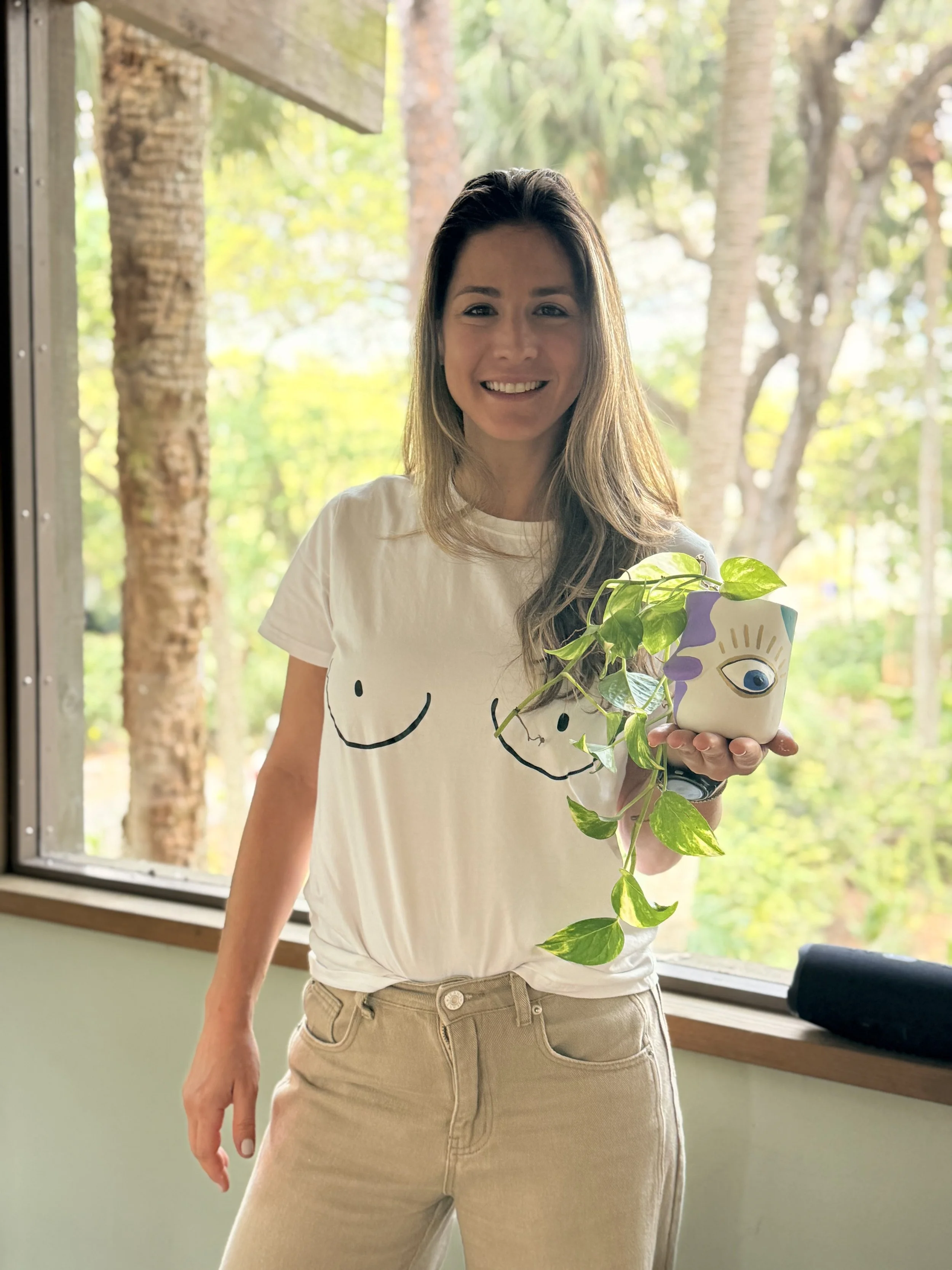 A woman smiling indoors near a window holding a plant in a colorful pot with a face design.