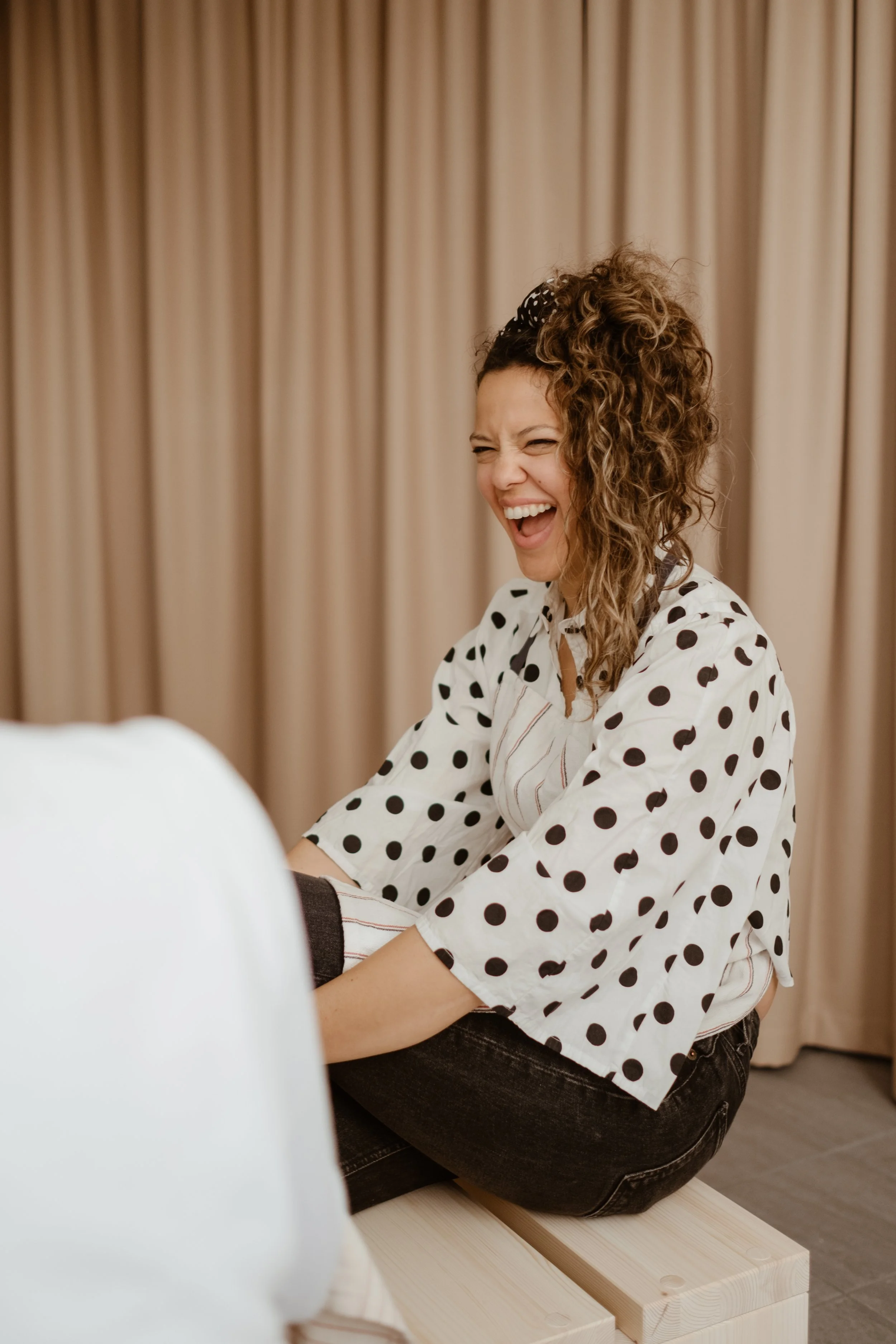 A woman with curly hair in a polka dot shirt laughing while sitting on a wooden bench with beige curtains in the background.