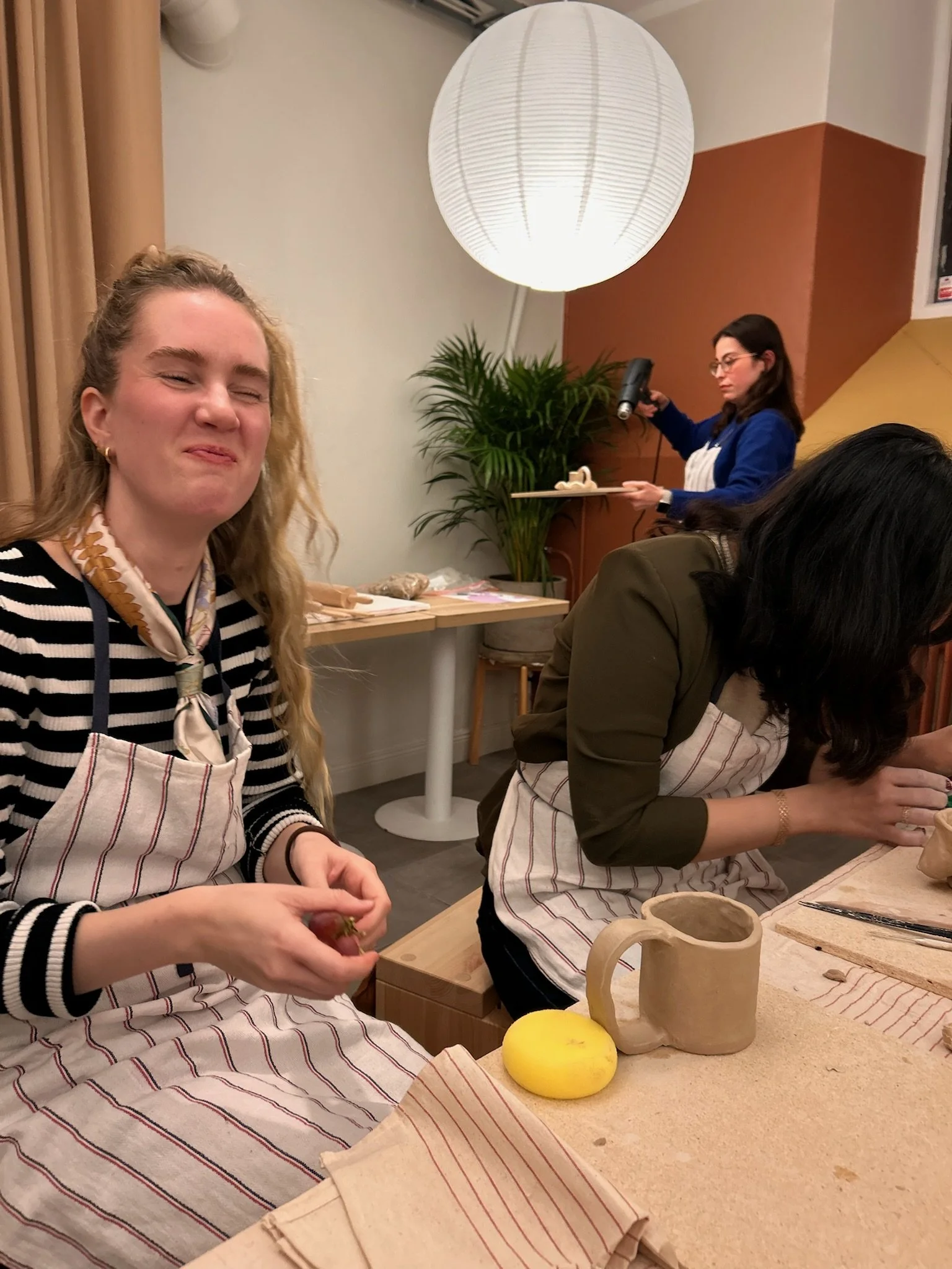 People participating in a pottery class, with ceramics and a mug on the table, a large round paper lantern overhead, and a woman in the background using a hairdryer.