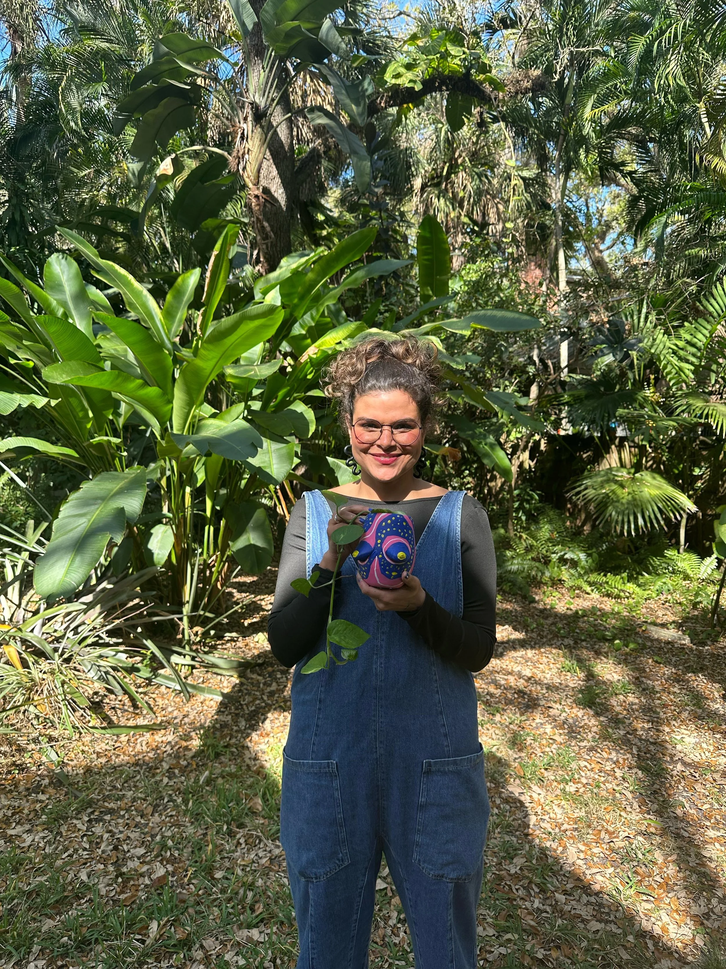 A woman with curly hair, glasses, and a smile, standing outdoors in a lush green garden, holding a colorful painted pumpkin.