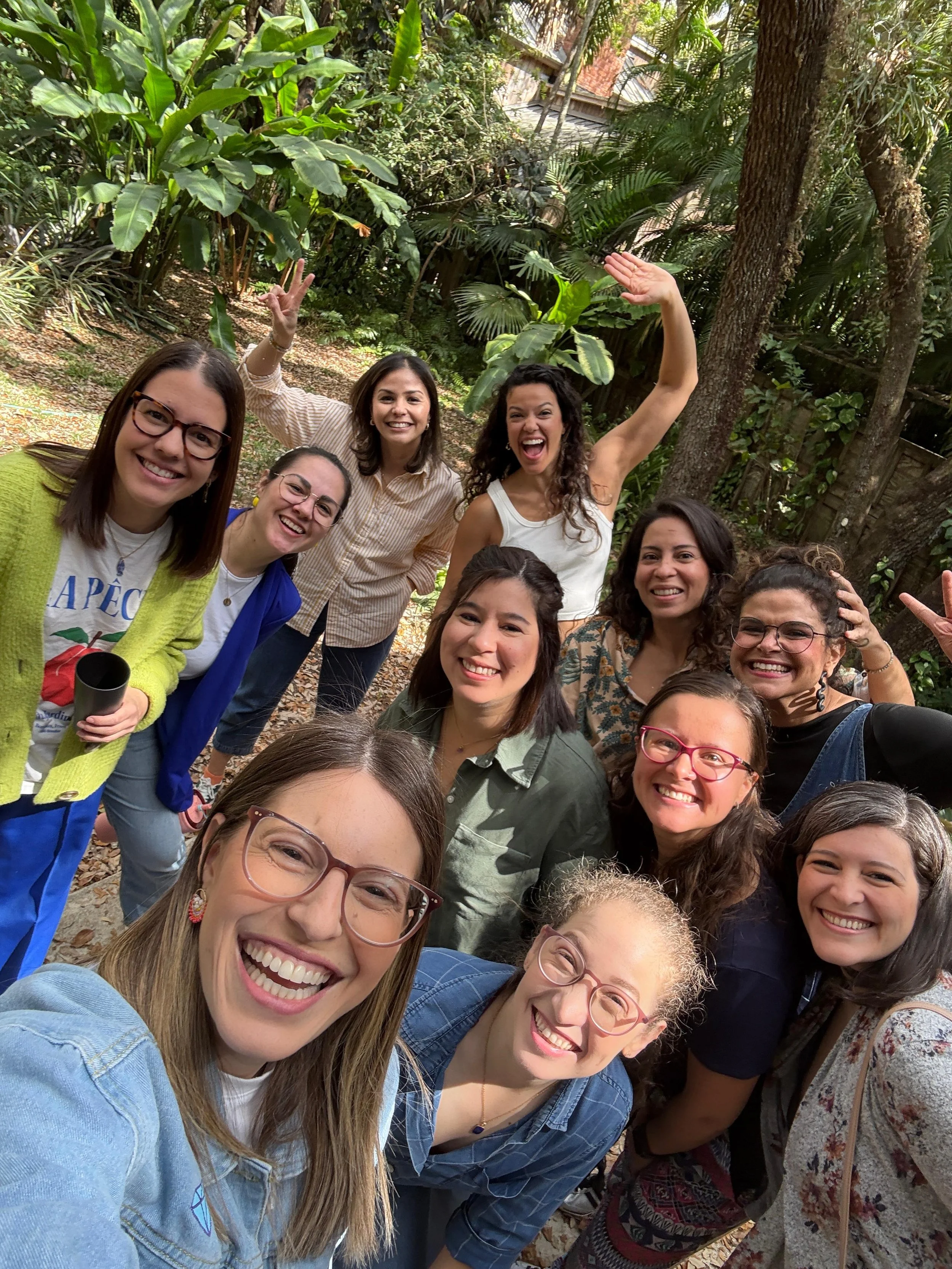 Group of women happily taking a selfie in a lush, green outdoor setting surrounded by trees and plants.