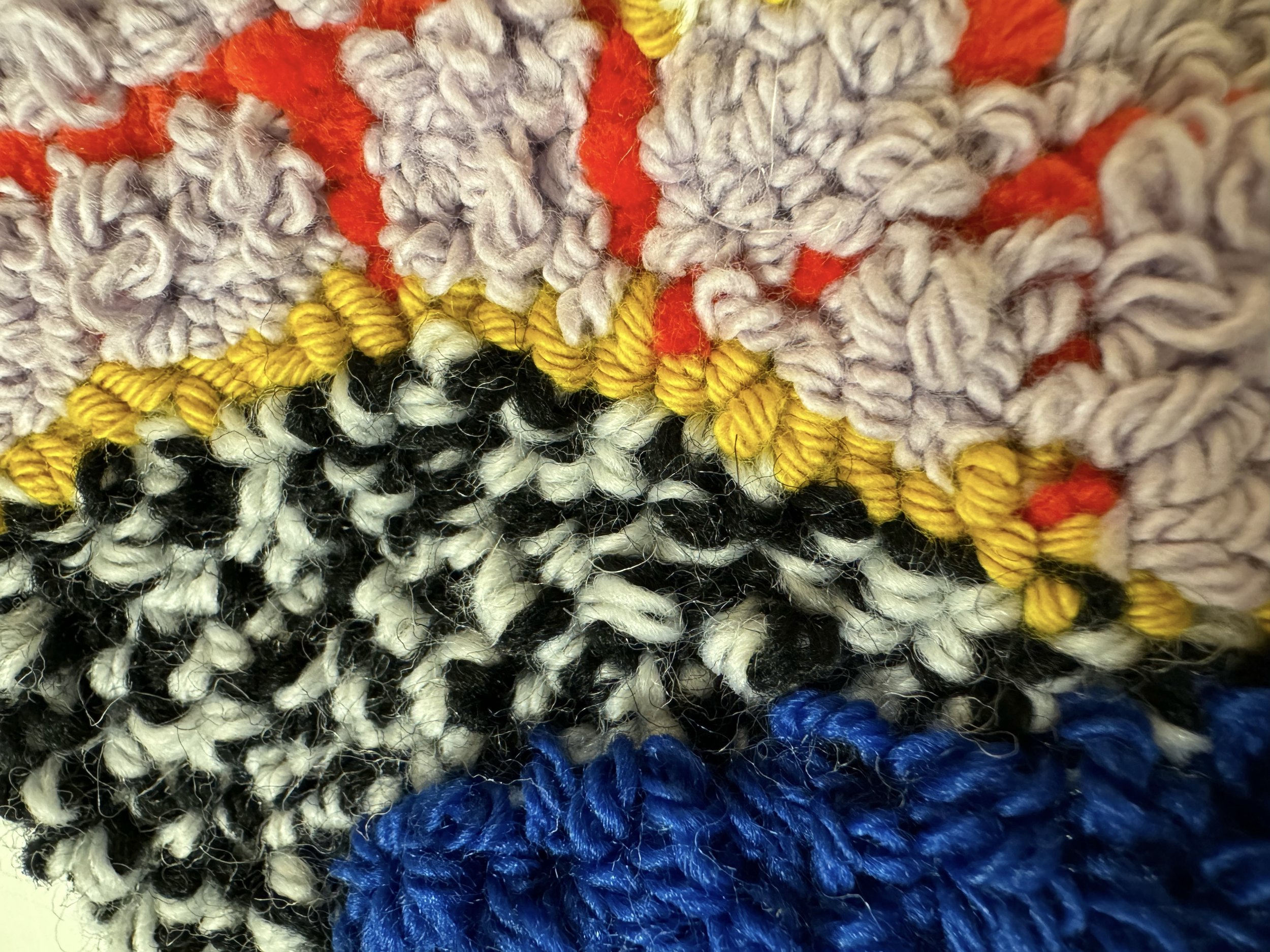 Close-up of a colorful, textured woven or knitted fabric with various shades including purple, yellow, black, white, red, and blue.
