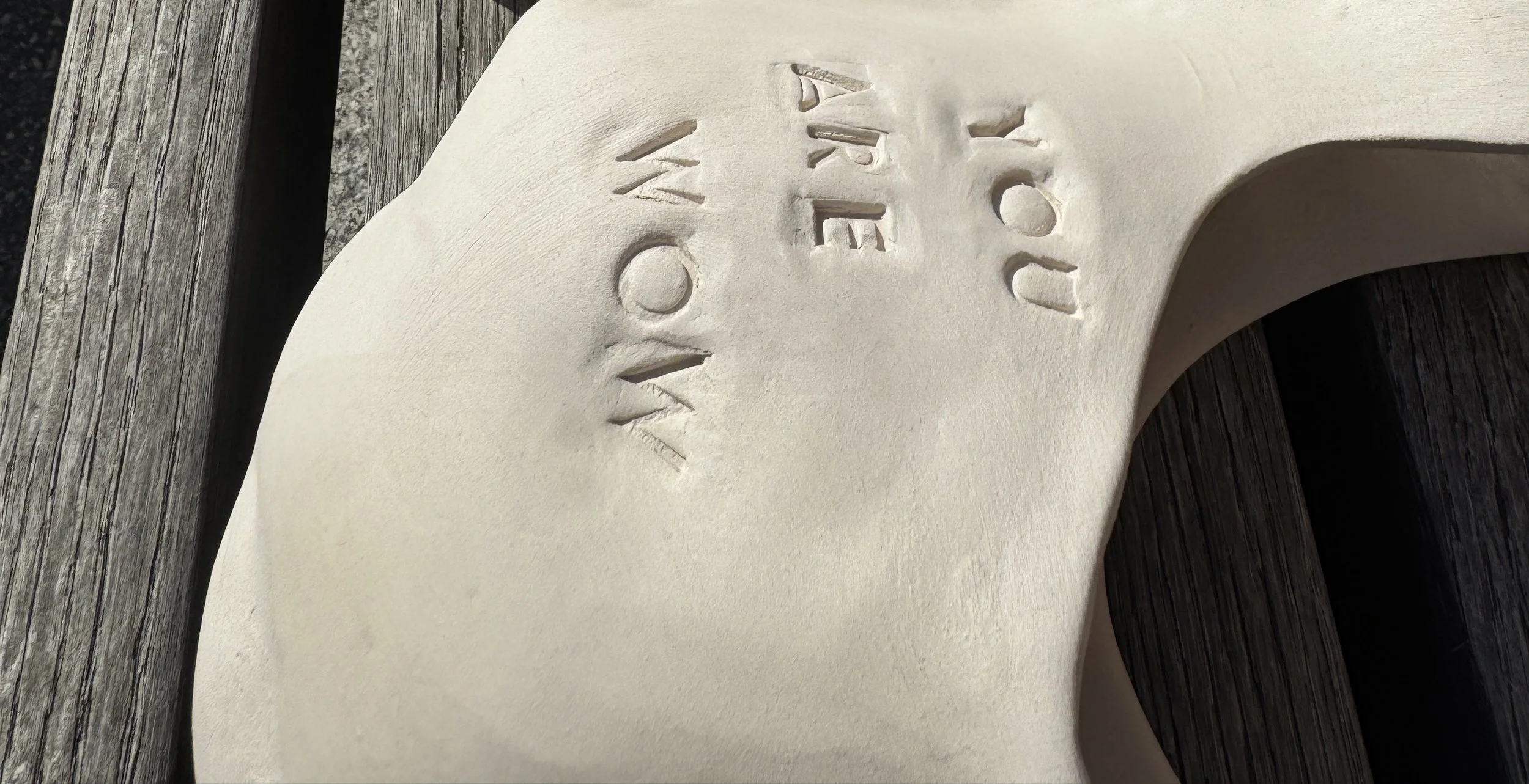 Close-up of a white ceramic chair with the words 'You Are Now' embossed on it, placed on a weathered wooden surface.