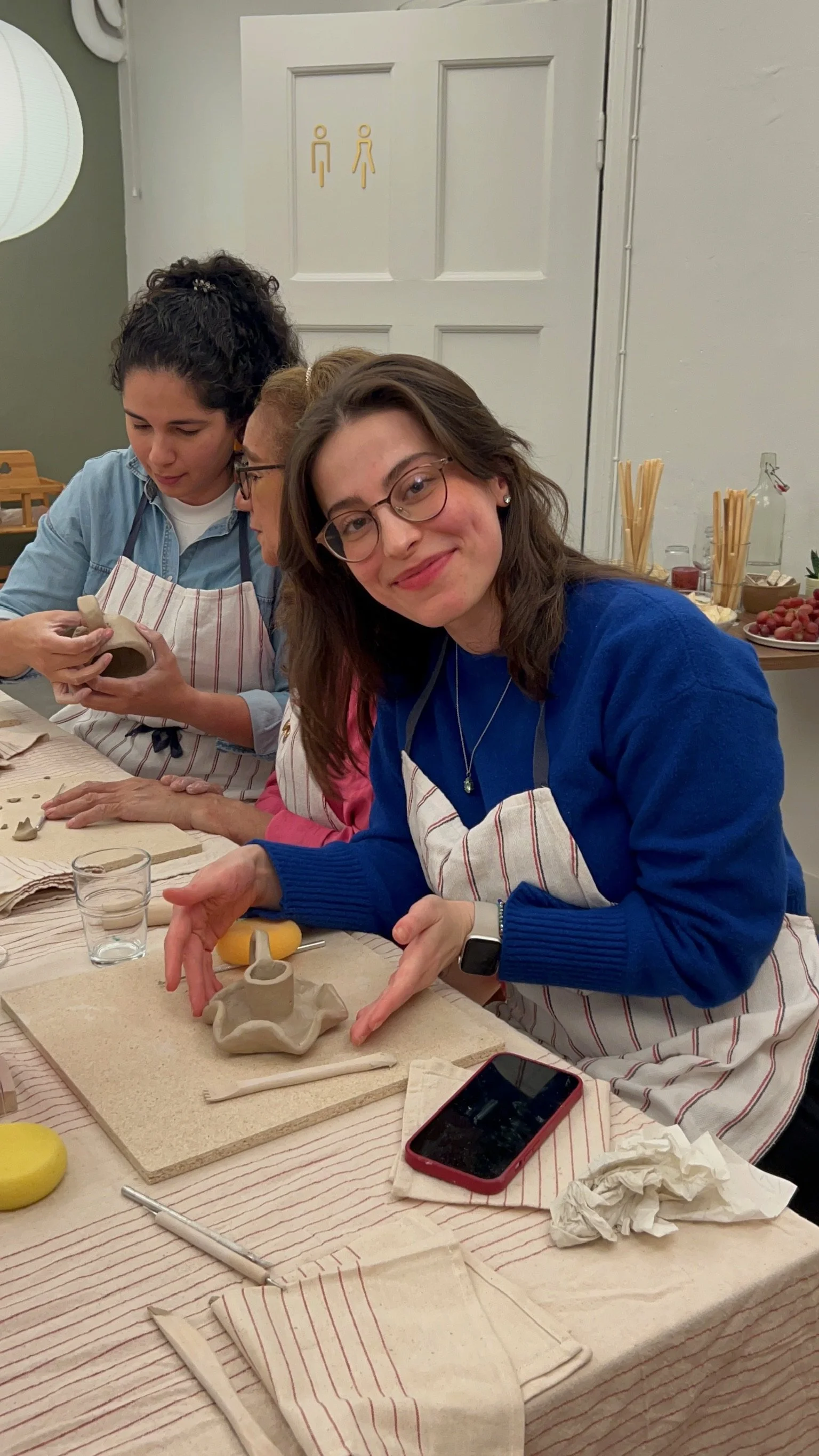 Woman with glasses smiling at camera, sitting at table with clay pottery. Other people working on clay projects in background. Art supplies and food on shelves behind.
