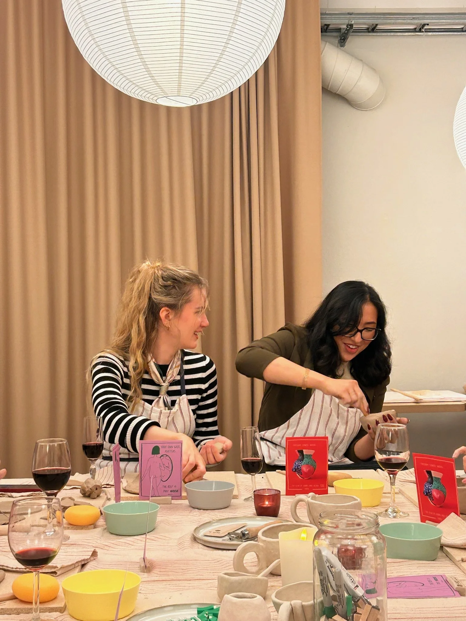 Two women smiling and engaging in a cooking class or workshop at a long table with bowls, wine glasses, and recipe cards, in a cozy indoor setting with beige curtains and hanging lantern lights.
