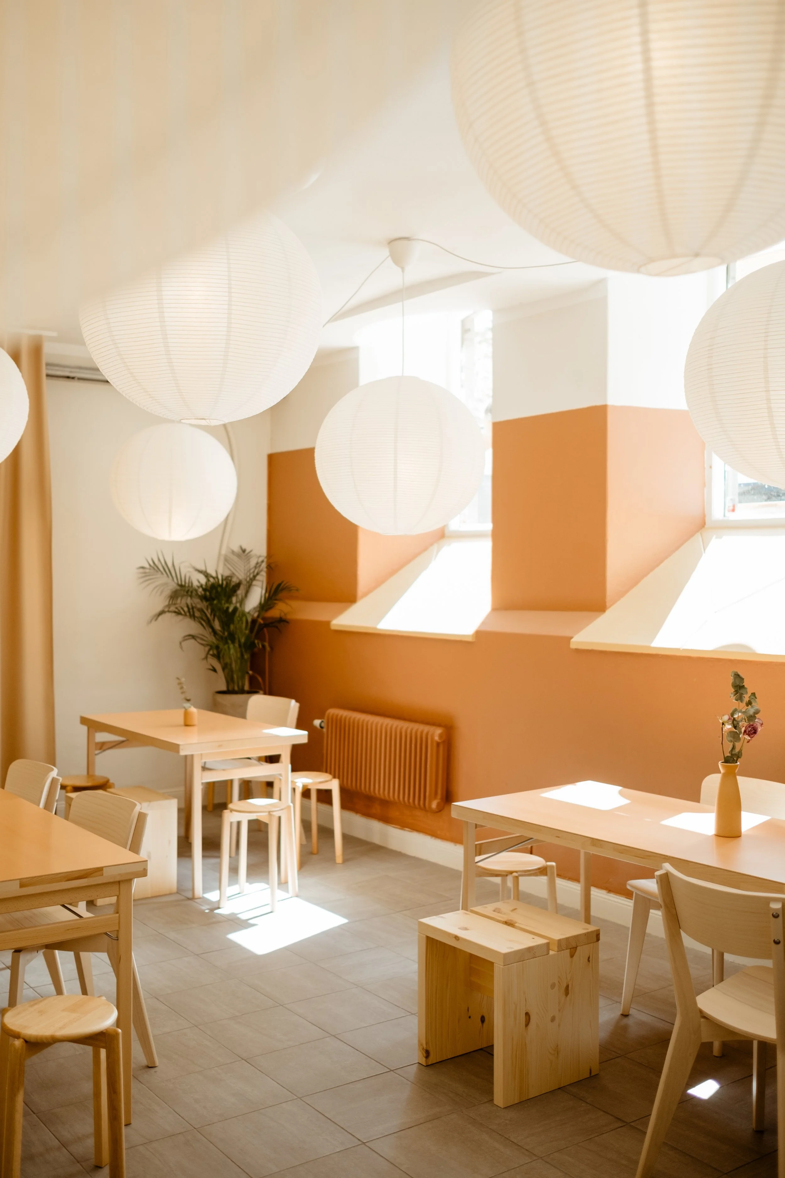 Bright, airy cafe interior with wooden tables, chairs, and stools, large paper lanterns hanging from the ceiling, peach-colored wall with windows, and a plant in the corner.