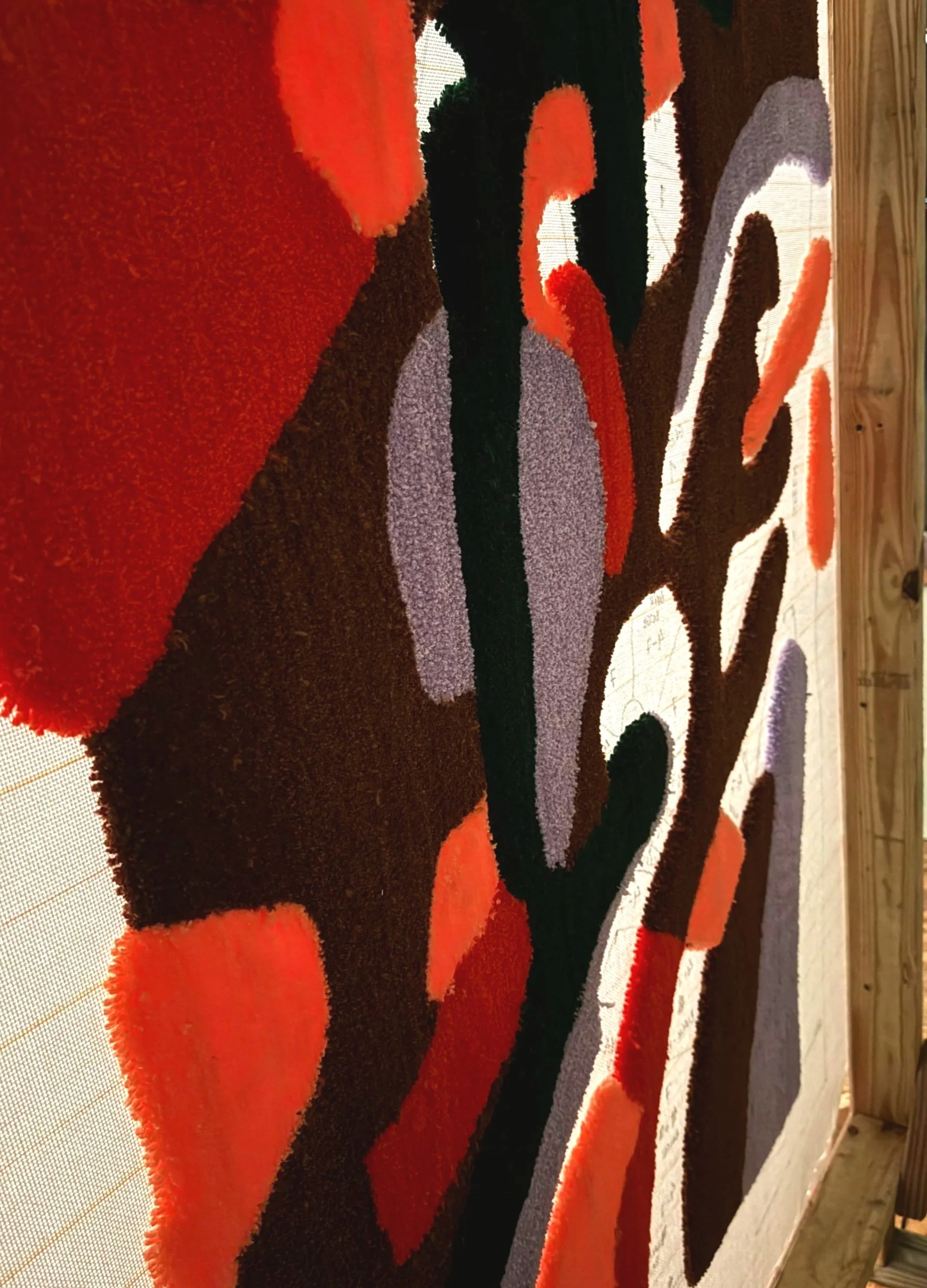 Close-up of a colorful, abstract textured rug with shapes in black, orange, purple, white, and red, placed on a wooden floor.