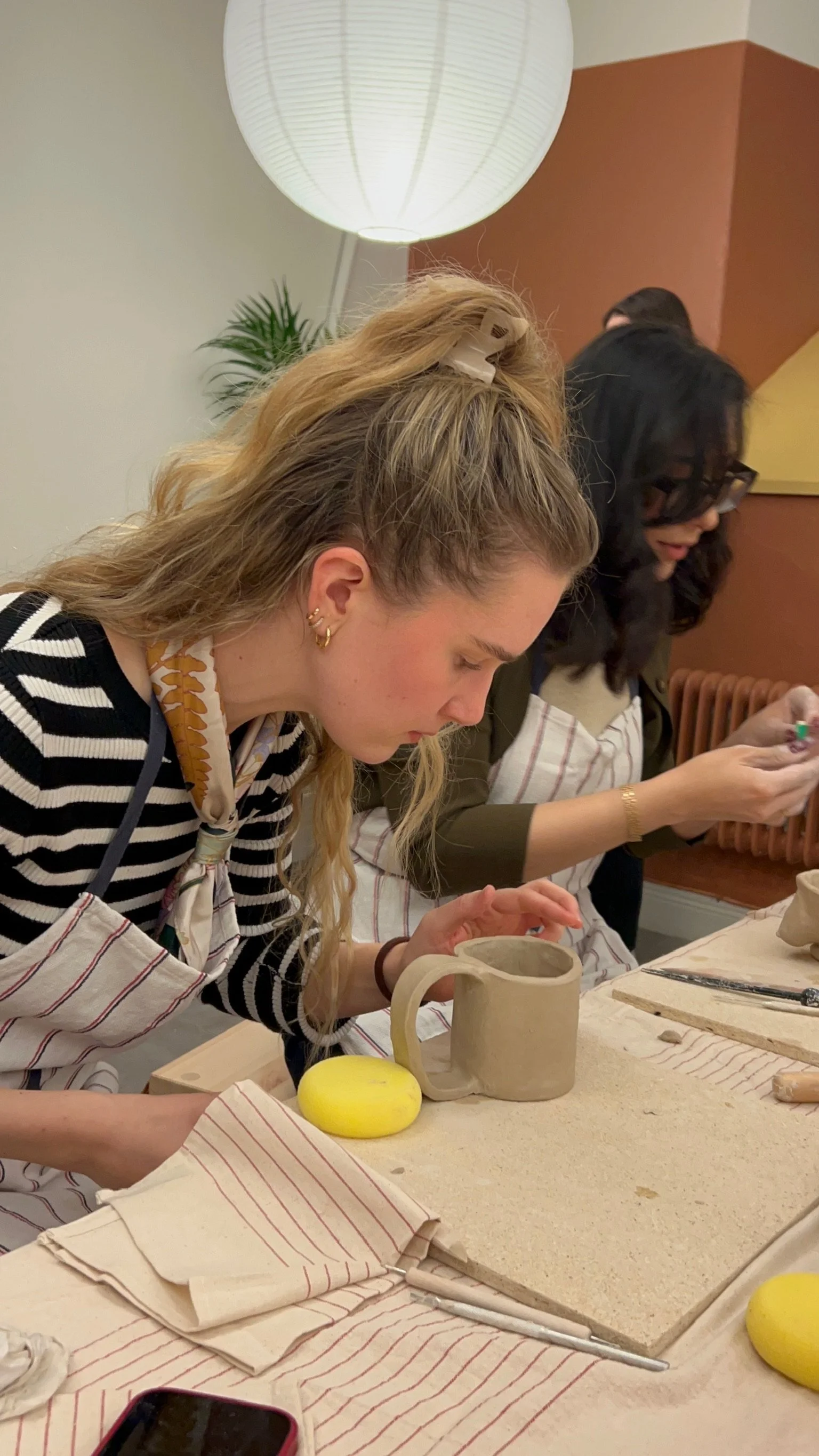 People participating in a pottery workshop, working on clay mugs, with pottery tools, in an indoor setting.