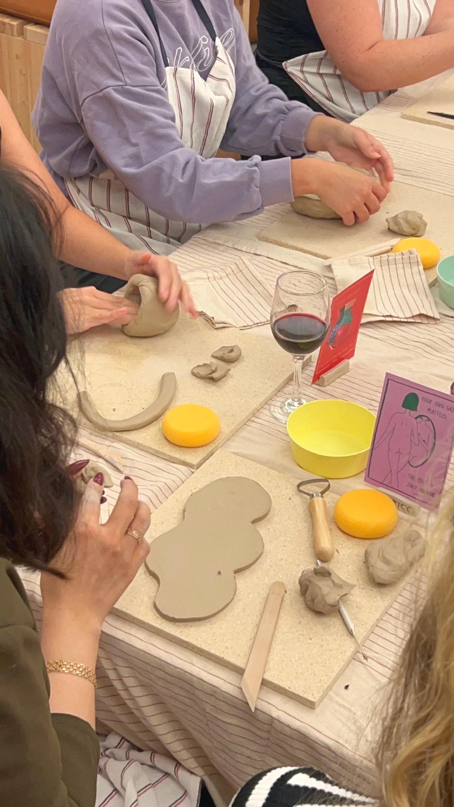Group of people gathered around a table, participating in a ceramic workshop. Various clay pieces, tools, and props are spread on the table, including a wine glass and colorful postcards.