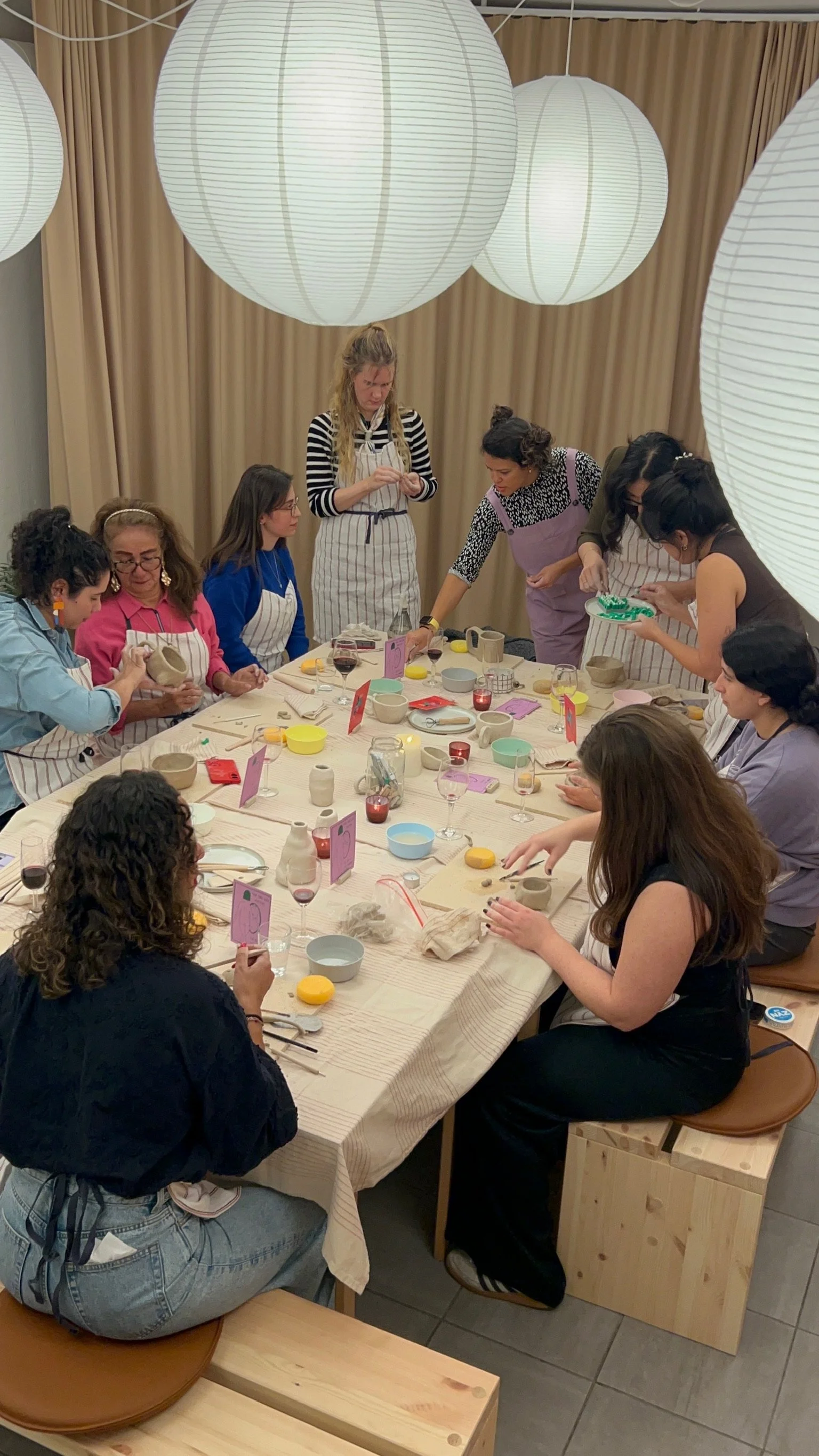 A group of women participating in a pottery workshop around a large table with pottery tools and unfinished clay pieces, illuminated by large paper lanterns hanging from the ceiling.