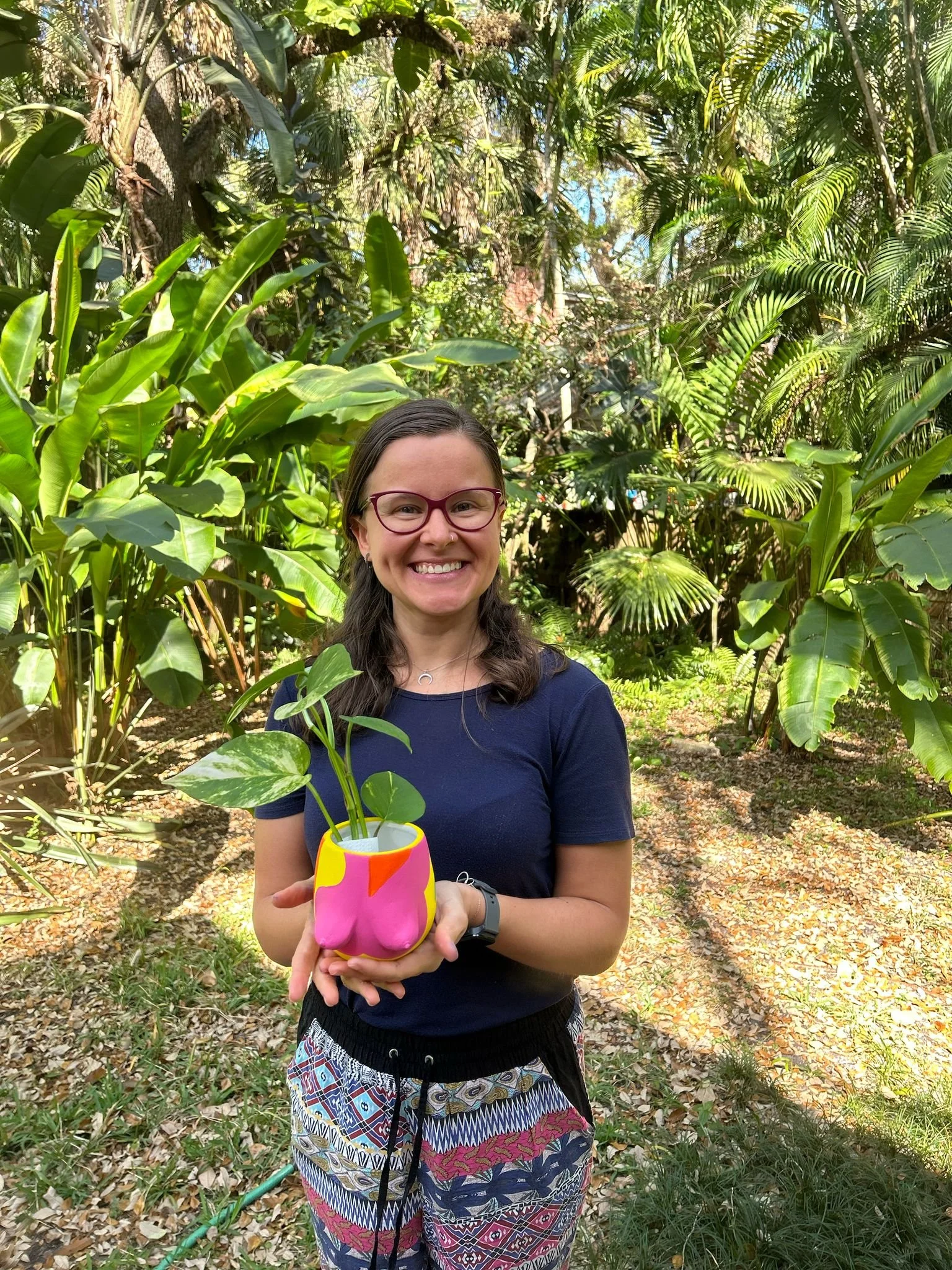 A woman with glasses smiling outdoors in a lush green garden, holding a colorful pink, yellow, and white pot with a plant in it.