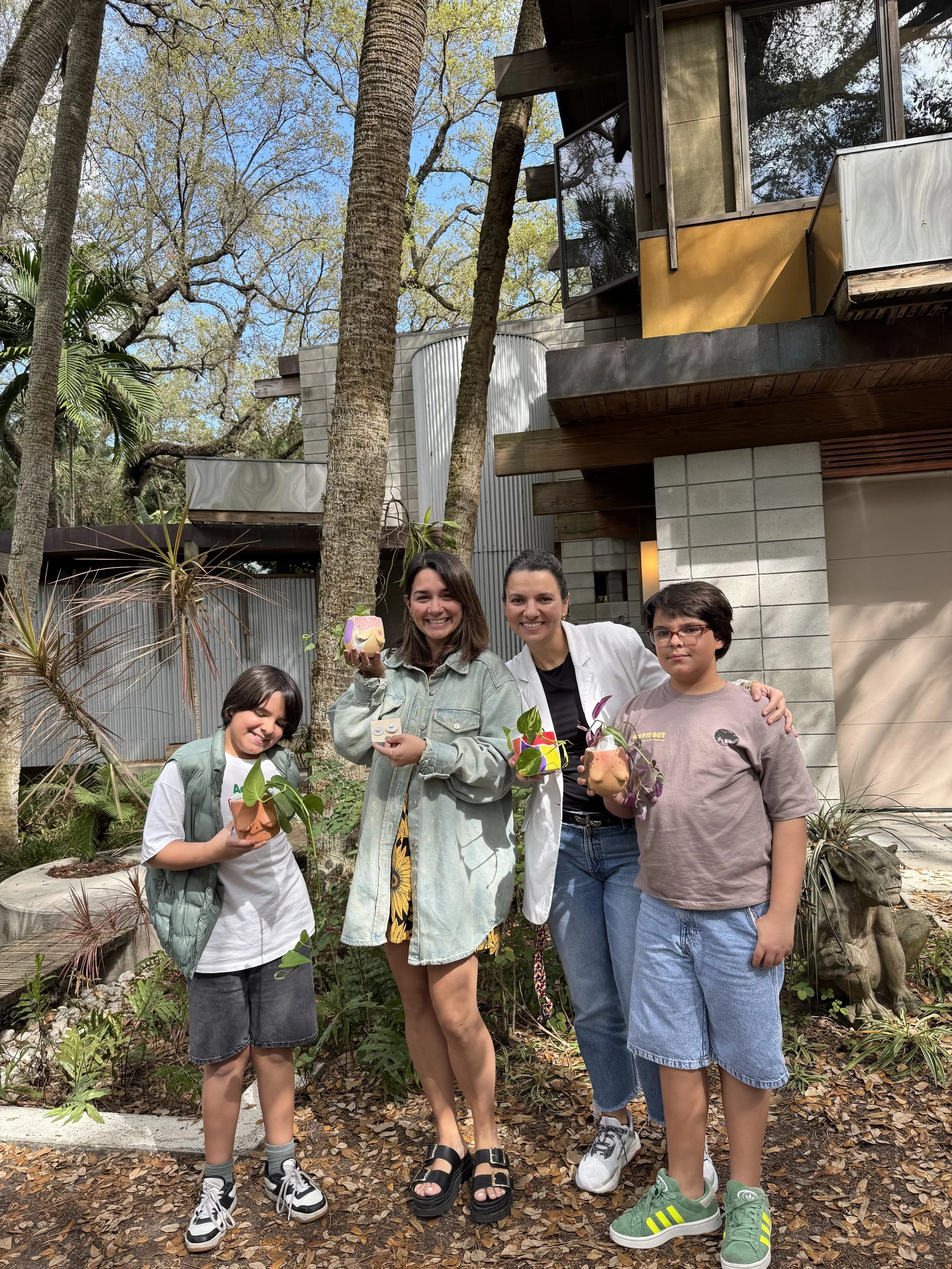 Four people, two children and two women, standing outdoors in front of a modern building and trees, holding colorful flower arrangements, smiling, with leaves and plants on the ground.