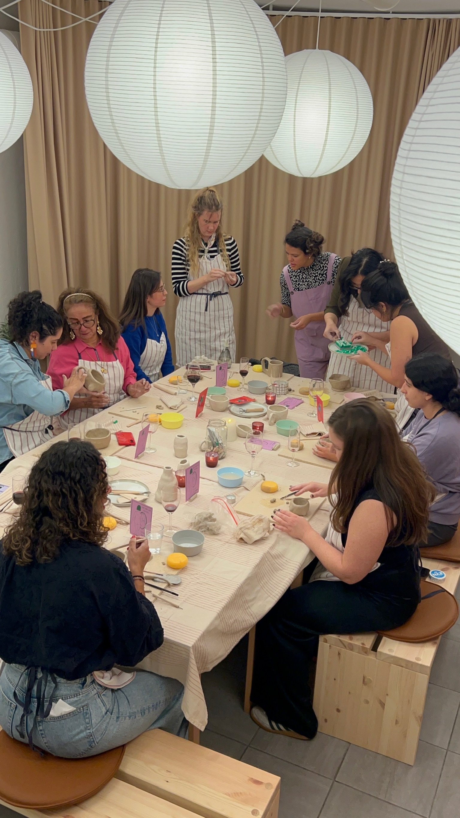 People gathered around a long table doing pottery painting in a decorated room with large paper lanterns hanging from the ceiling.