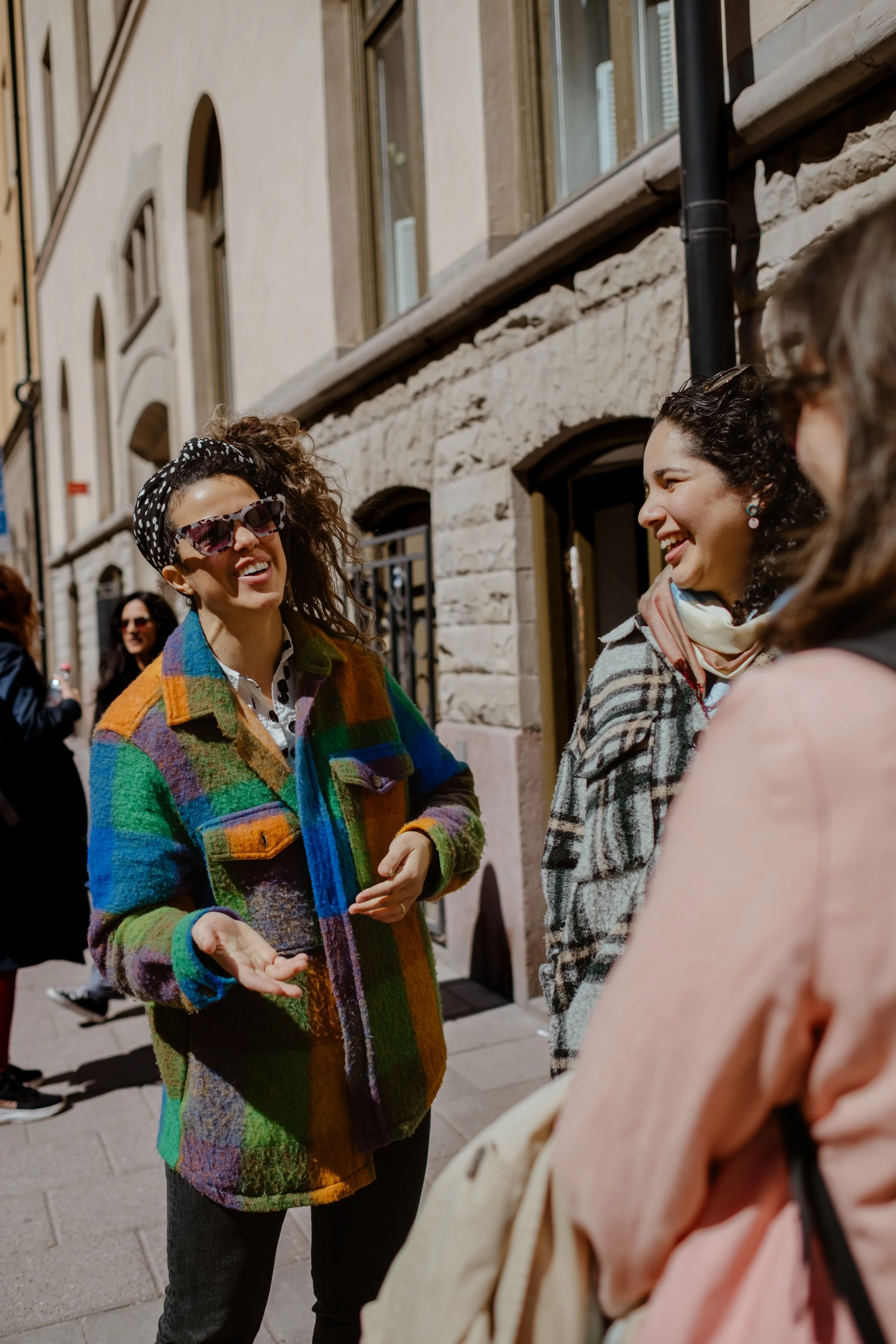 Three women are standing on a city sidewalk sharing a laugh and conversation. One woman wears sunglasses and a colorful rainbow patchwork jacket, another wears a plaid coat with a scarf, and the third woman is partially visible in the foreground with a light pink jacket.