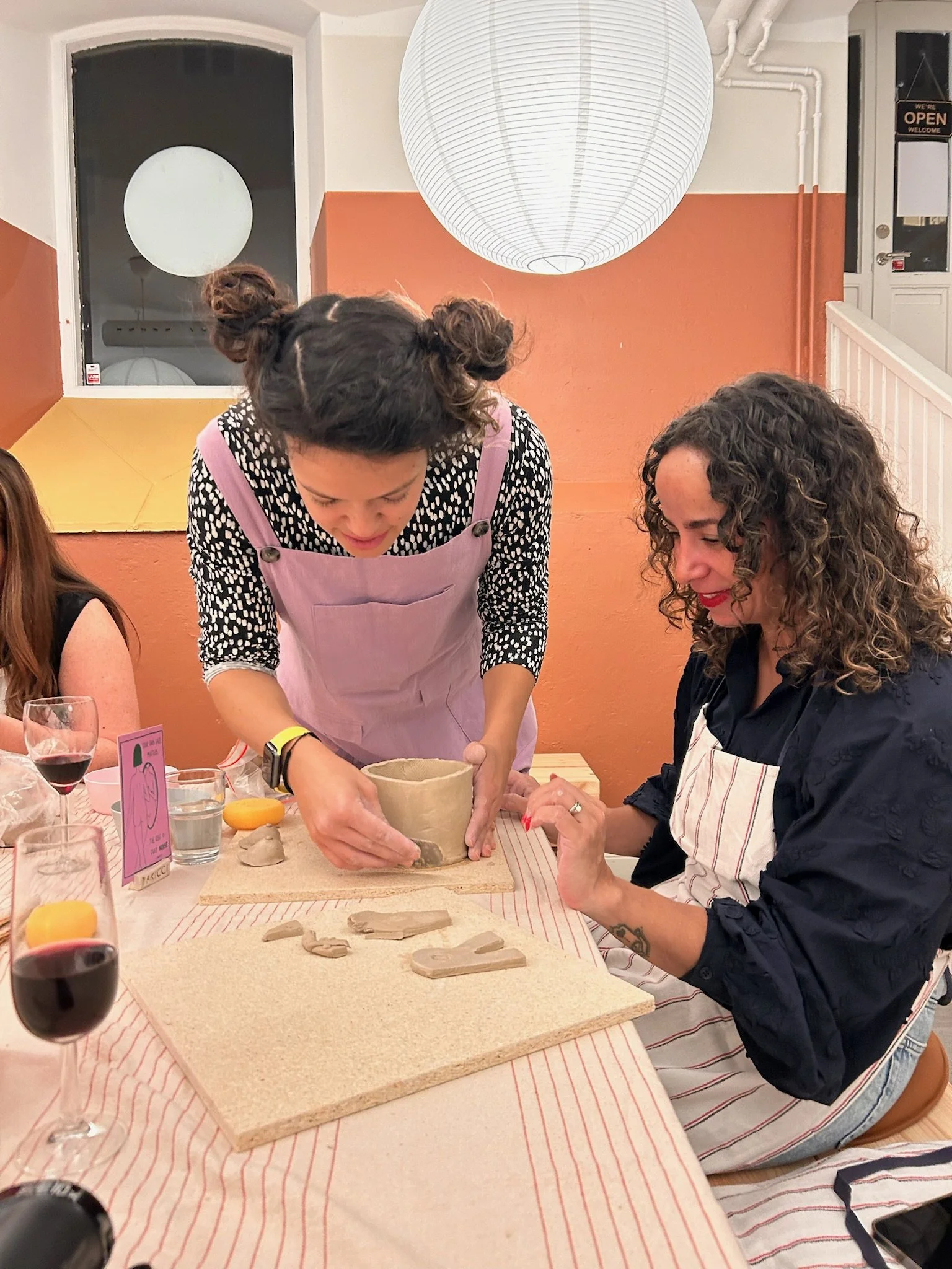 A woman with curly hair and a black shirt sits at a table while a woman with styled hair and a pink apron is working on a ceramic project. The table has wine glasses, a water glass, and a card with a pumpkin illustration. The room has warm lighting a