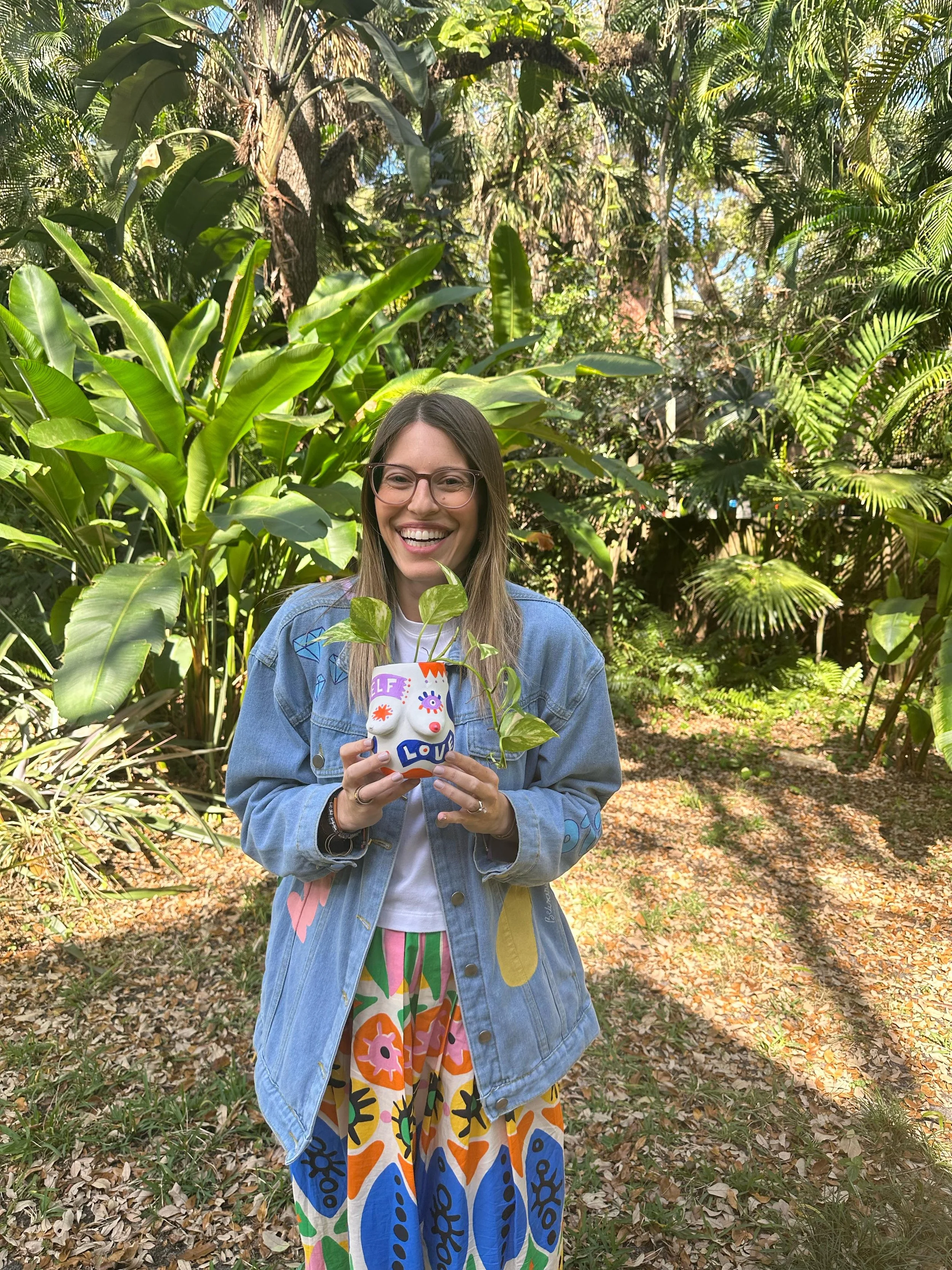 A woman standing in a lush, green garden, holding a colorful, handmade ceramic plant pot with a plant growing from it. She is smiling, wearing glasses, a denim jacket with colorful patches, a white shirt, and a vibrant, patterned skirt.