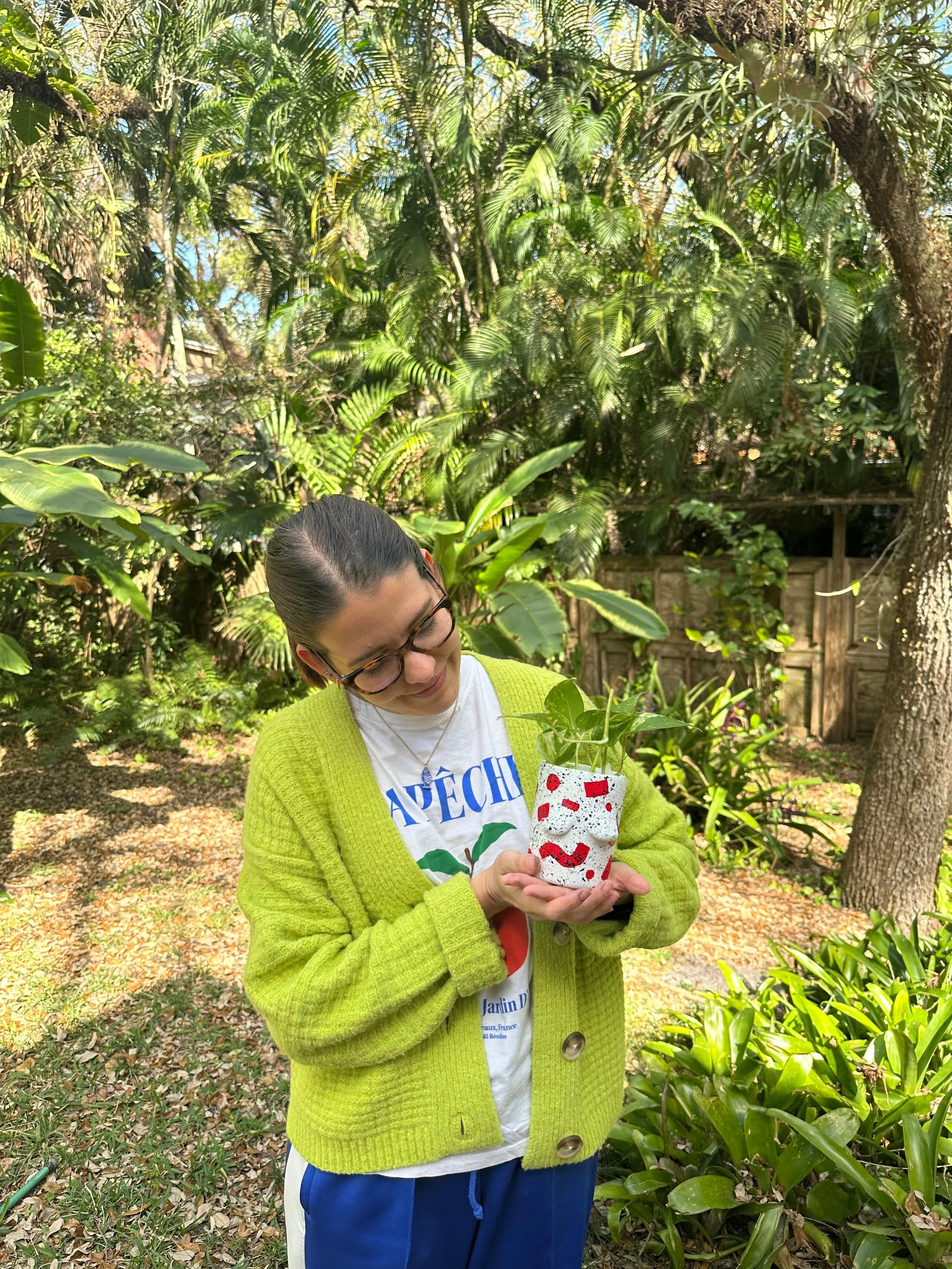 Young woman with glasses holding a small, decorated plant pot with green foliage, standing in a lush garden with trees and plants.